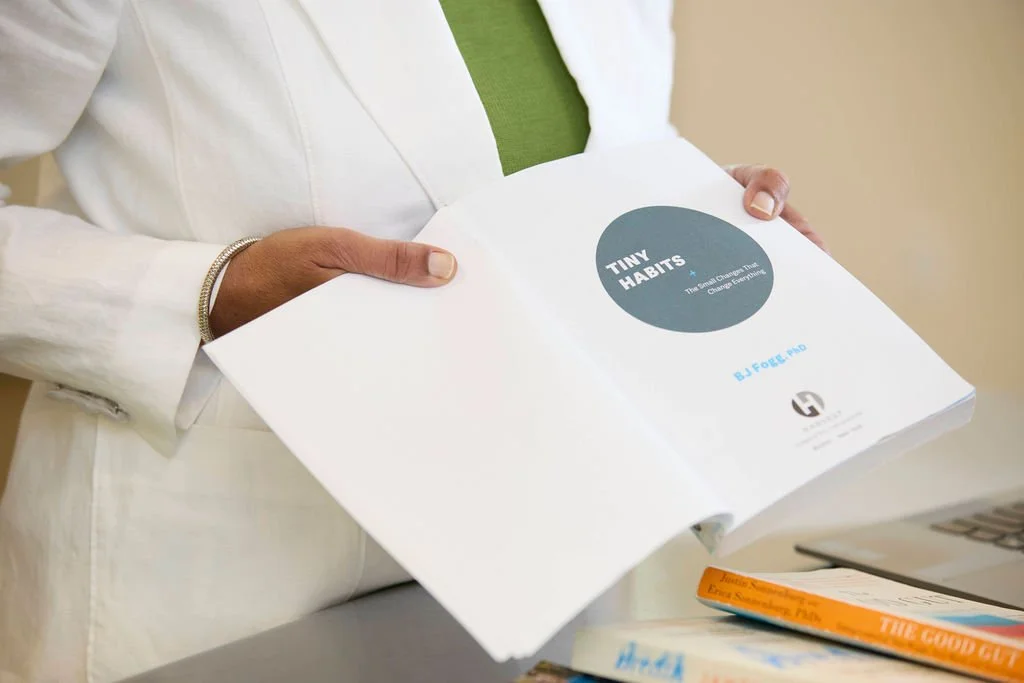 Person holding an open booklet titled 'Tiny Habits' with a green shirt visible underneath a white jacket.