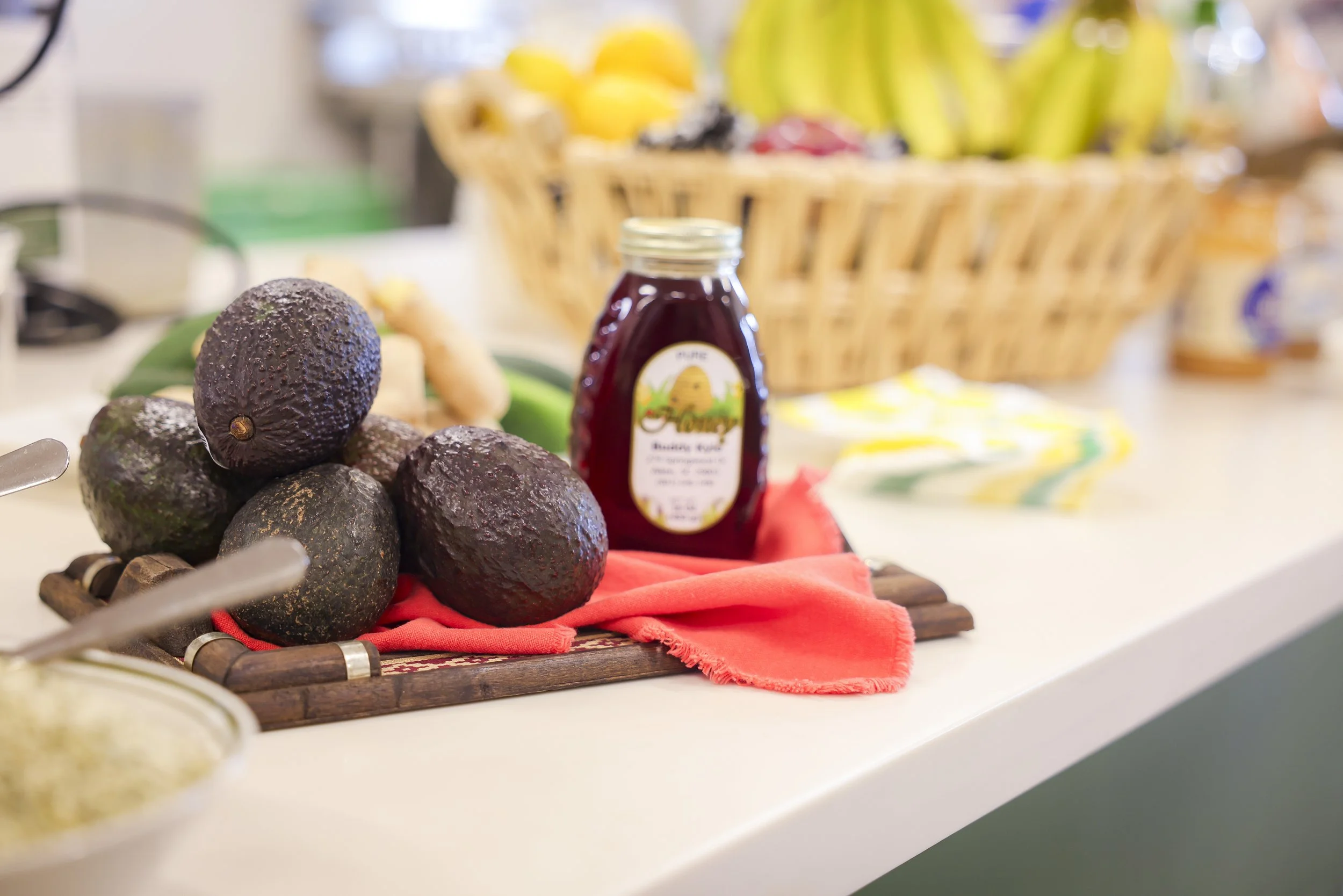 Avocados and jar of jam on a kitchen counter with a basket of bananas and other fruits in the background.