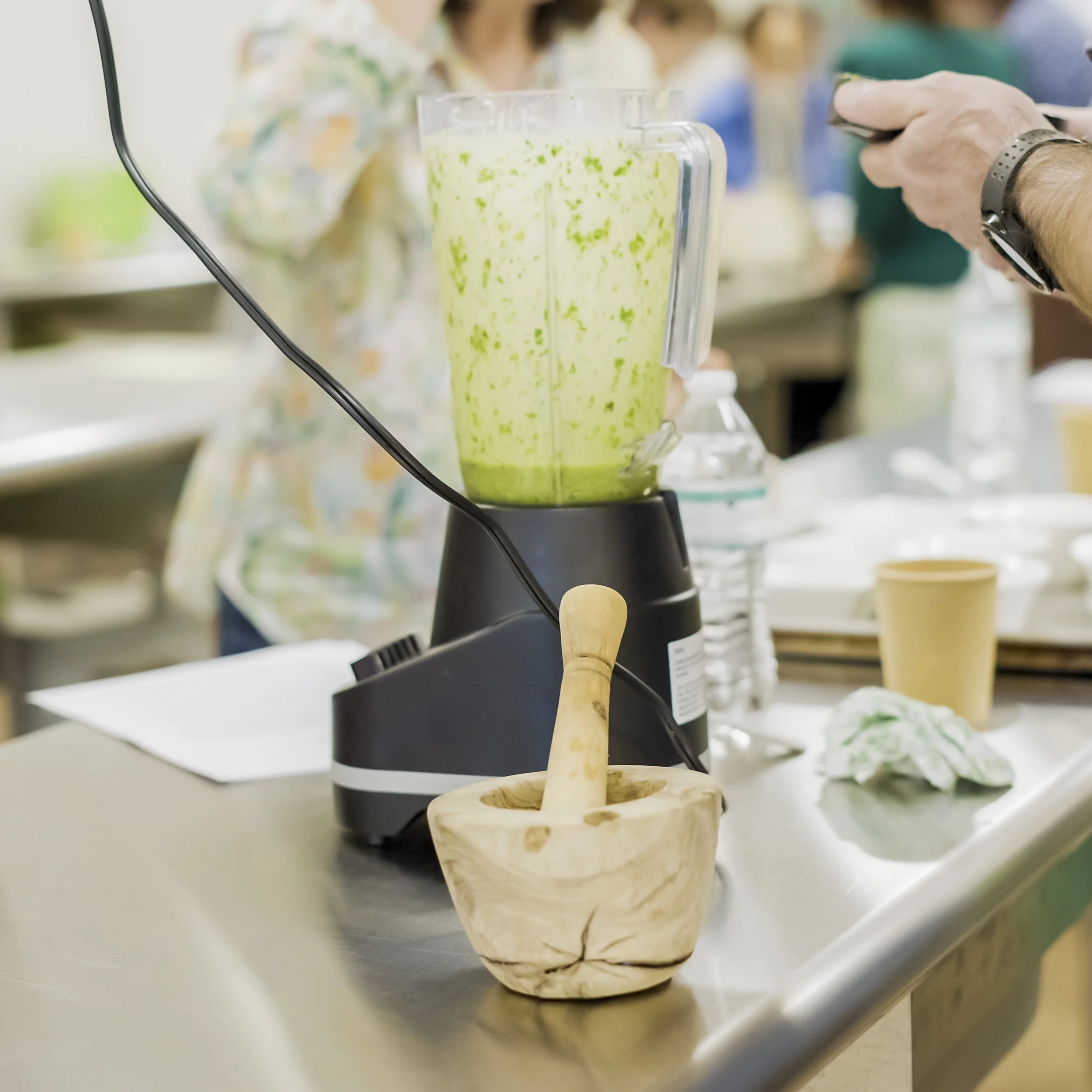 A blender with a green smoothie mixture inside on a stainless steel countertop, with a wooden mortar and pestle in front, and a person in the background holding a smartphone.
