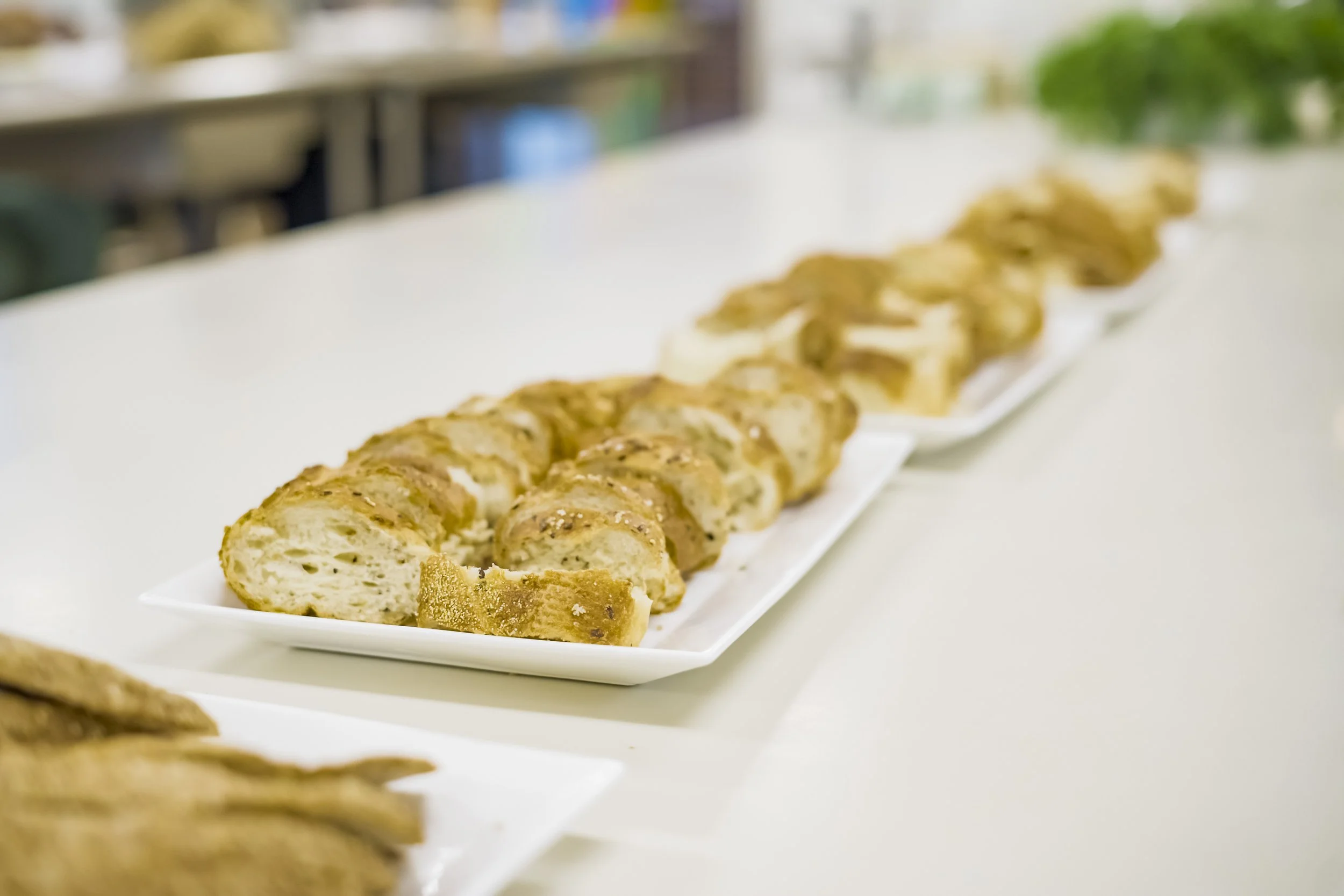 Close-up of sliced bread or bread rolls on a white rectangular plate, with other similar bread items in the background on a white surface.