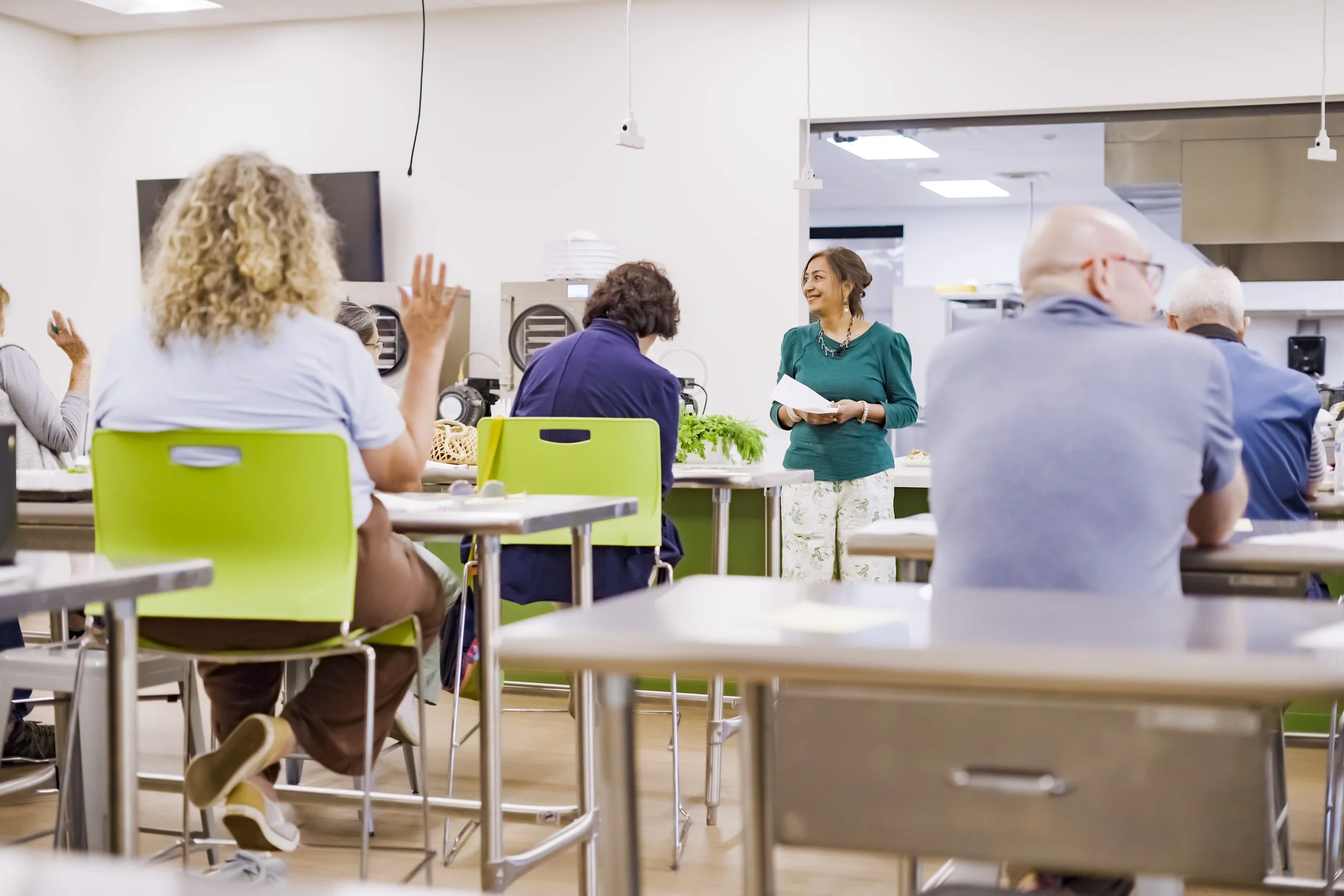 A woman in a green top and white patterned pants speaking to a group of people in a classroom or seminar setting, with attendees seated at tables and one person raising their hand.