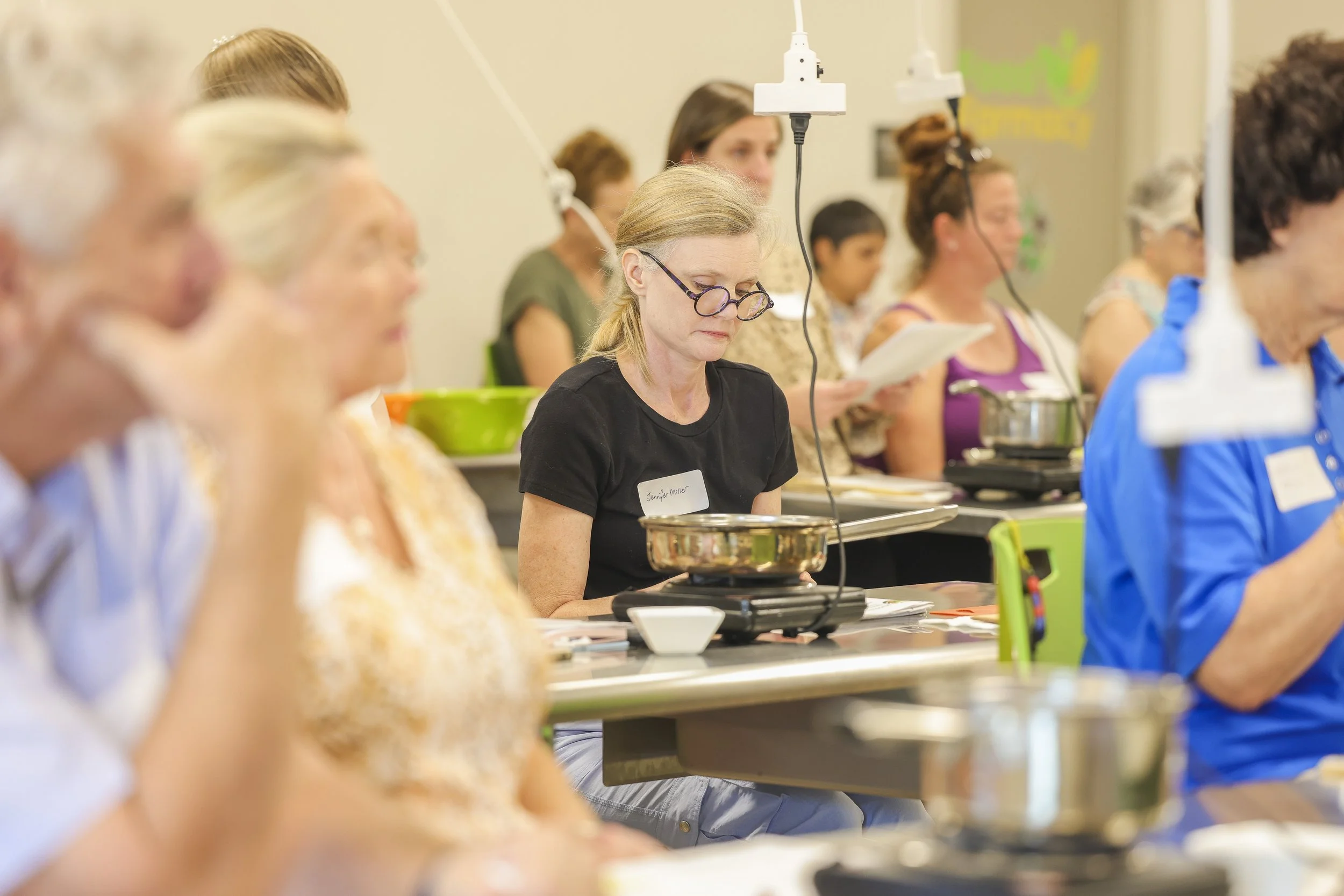 A group of people attending a cooking or food tasting class, seated at tables with cooking equipment, with a woman in black focused on her tasting.