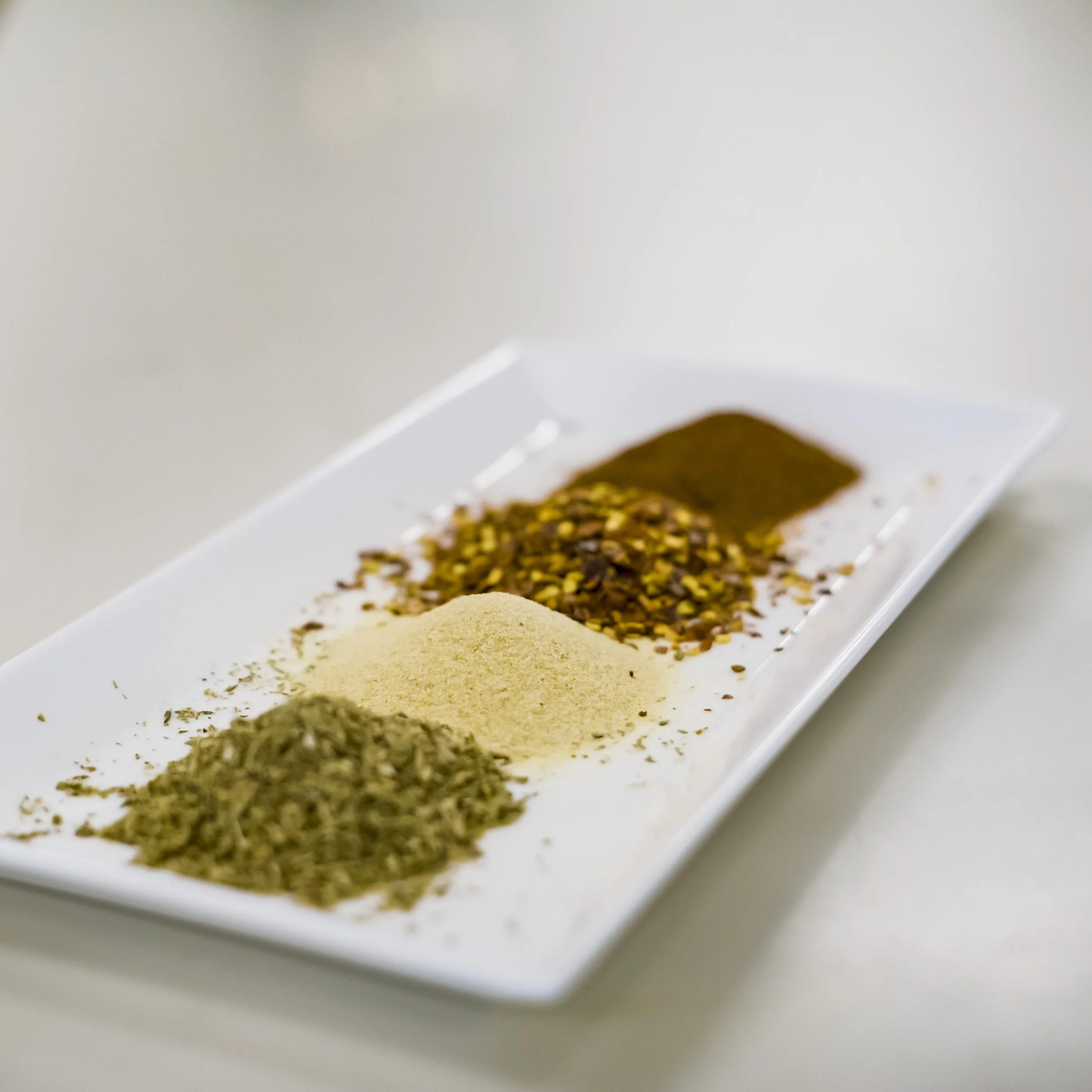 Four different spices on a white rectangular plate, arranged in rows, with various textures and colors.