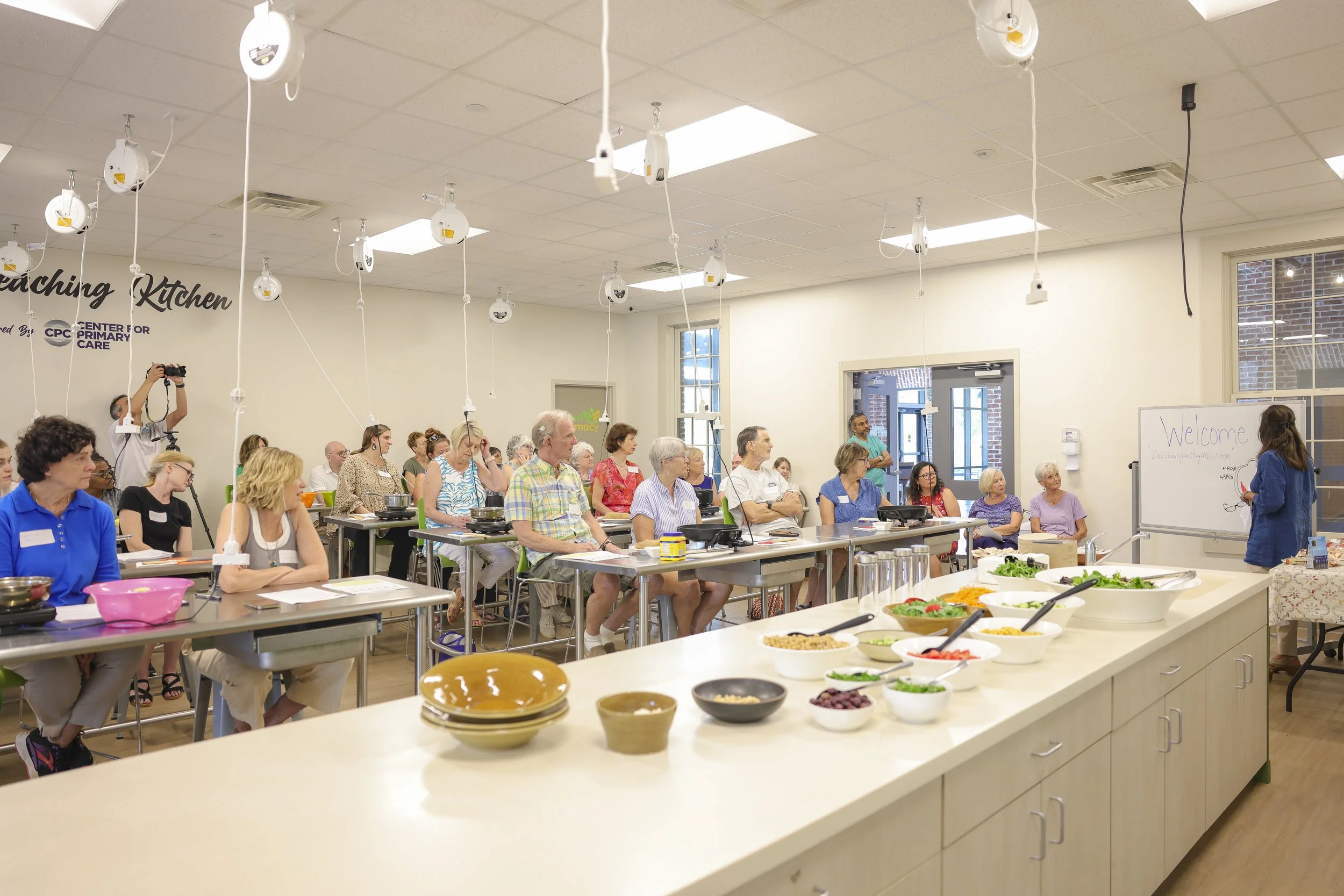 A cooking class or demonstration in a large bright room with a white counter in the foreground, bowls of salad ingredients, and a group of diverse adults seated at tables watching a woman instructor at the front near a whiteboard. The room has ceilin