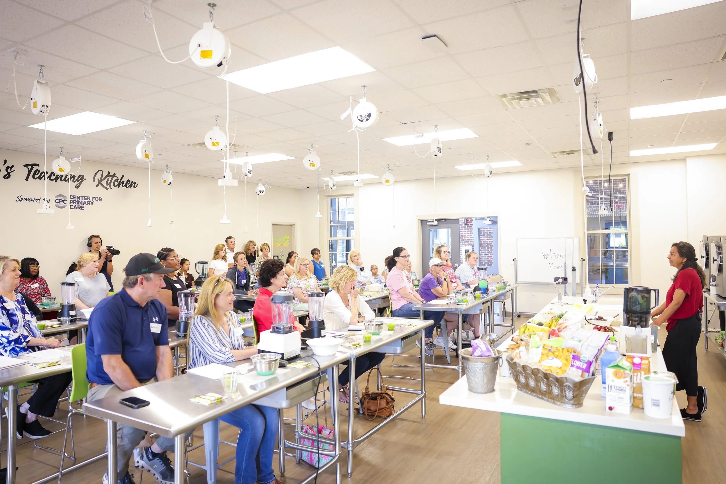 A cooking class or workshop in a brightly lit room with an instructor standing at the front, teaching a group of adults seated at tables with cooking ingredients and appliances. There are multiple ceiling-mounted cameras and a whiteboard that says "W