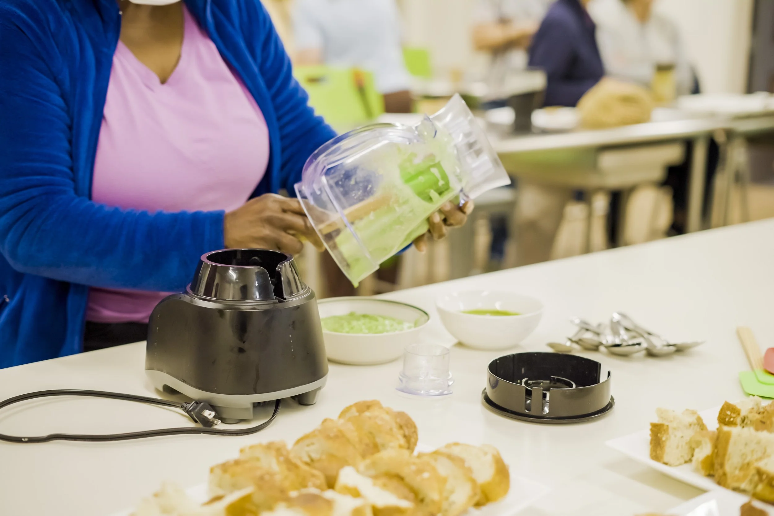 A person in a blue jacket and pink shirt preparing food at a table with a bread toaster, bowls of green spread, and sliced bread, in a communal kitchen or cafeteria setting.