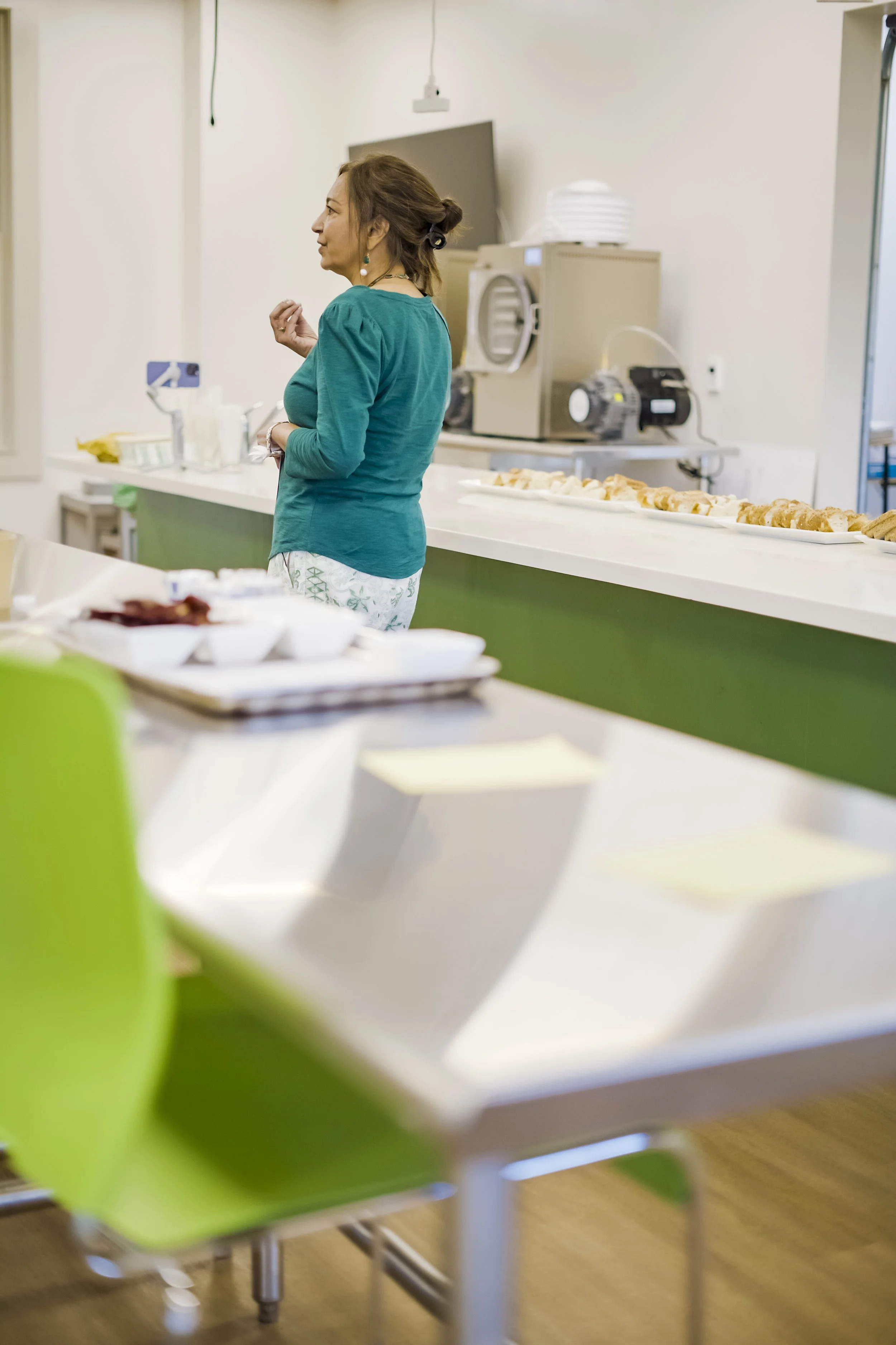 A woman in a teal shirt and patterned pants standing in a cafeteria with food trays and kitchen equipment in the background.