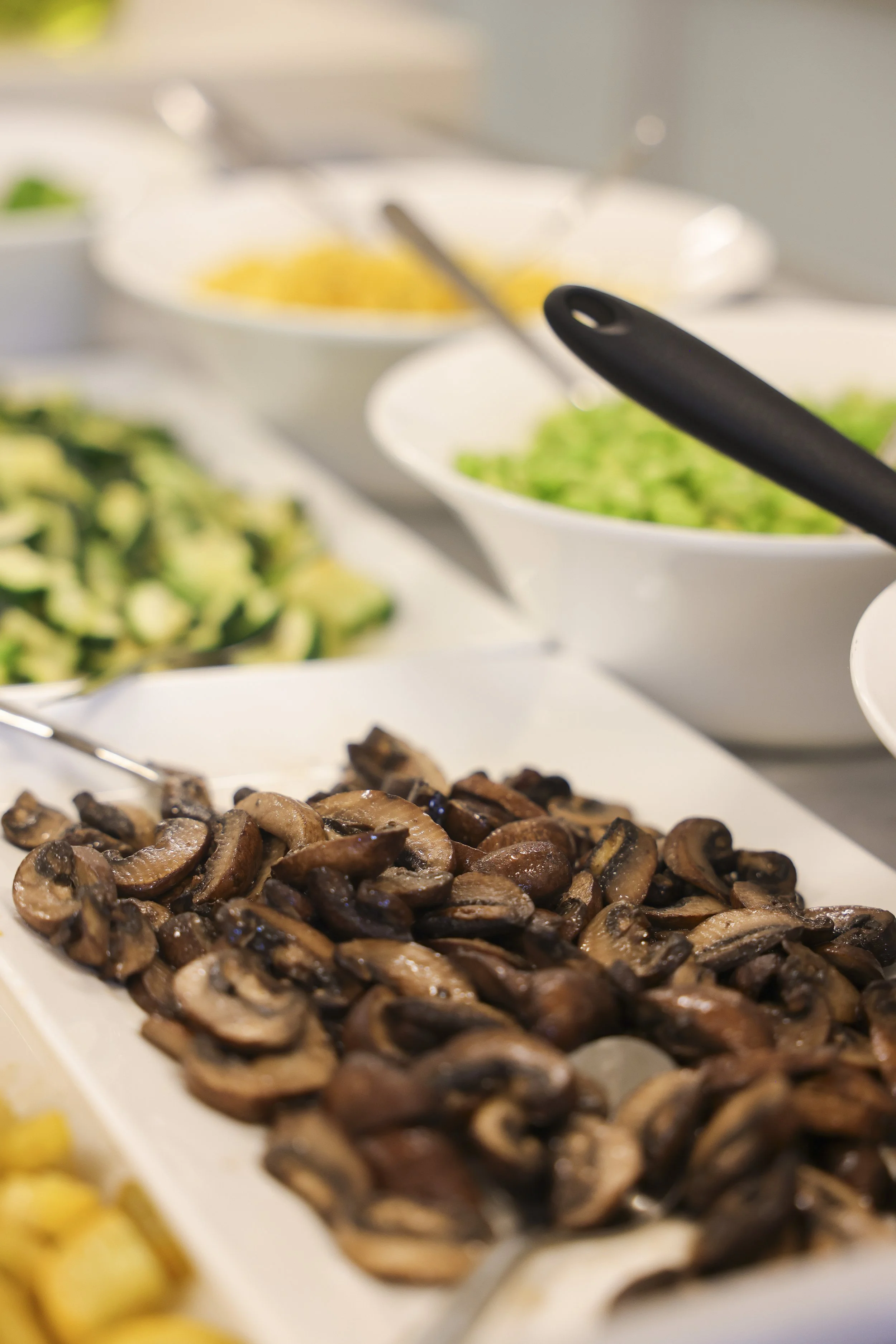 Close-up of a buffet with sliced mushrooms on a white serving platter, with bowls of chopped greens and other salad toppings in the background.