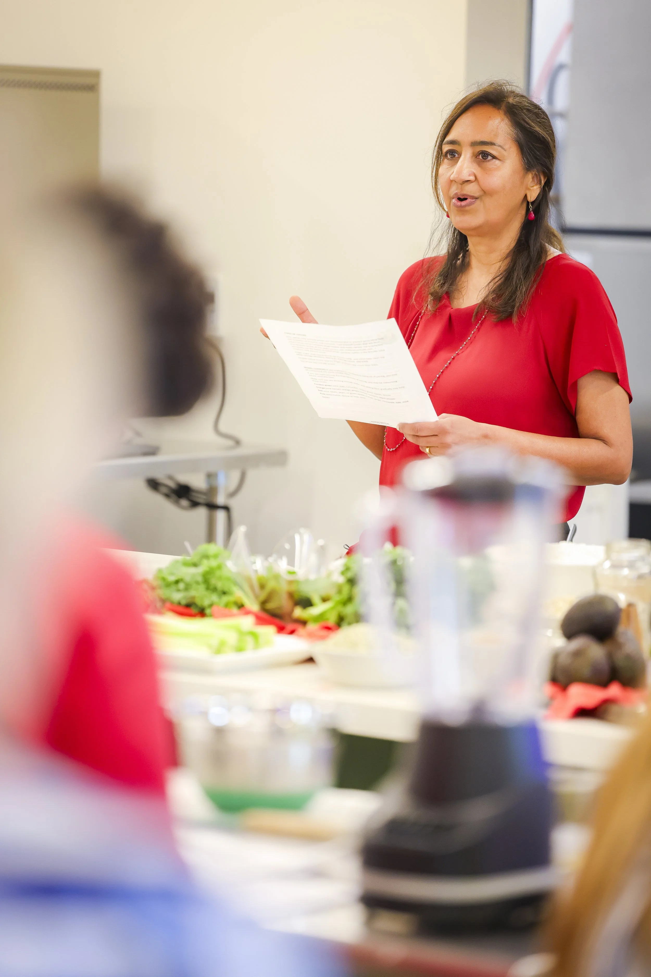 A woman in a red top holding a paper, speaking in front of a group during a presentation or meeting. There are various food items on the table in front of her.