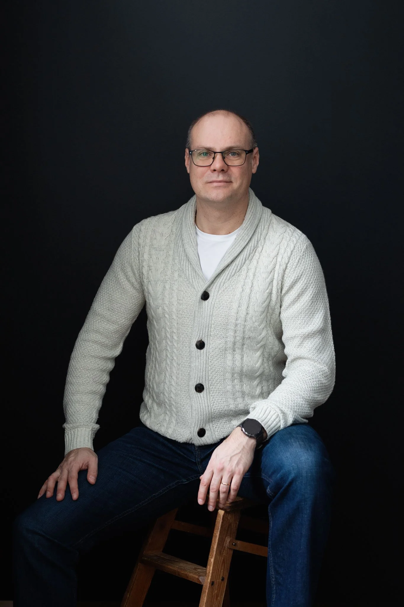 A man with glasses wearing a white cable-knit cardigan over a white shirt, sitting on a wooden stool against a black background.