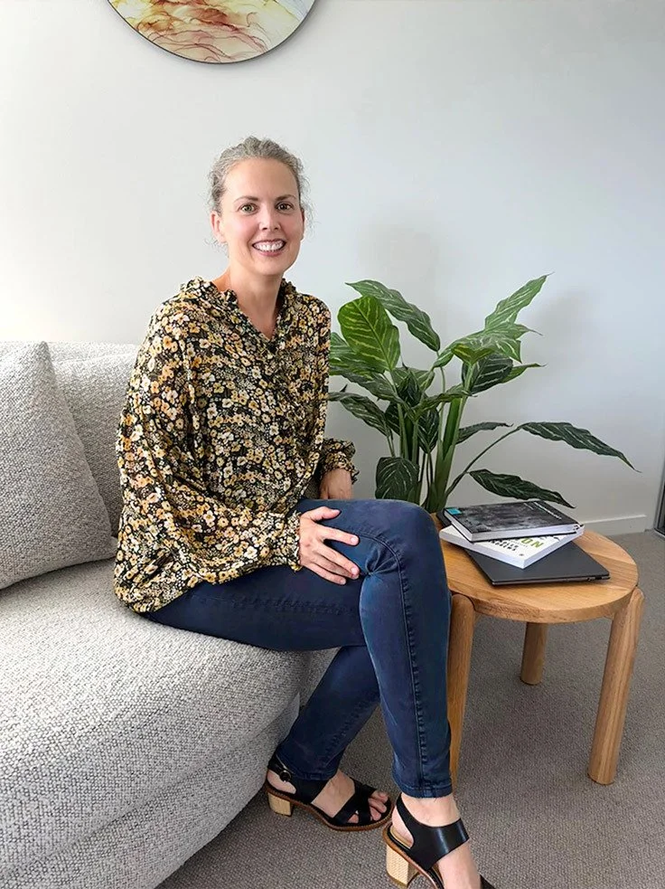Smiling Centred Wellness therapist sitting on a grey sofa next to a wooden table with books and a potted plant.