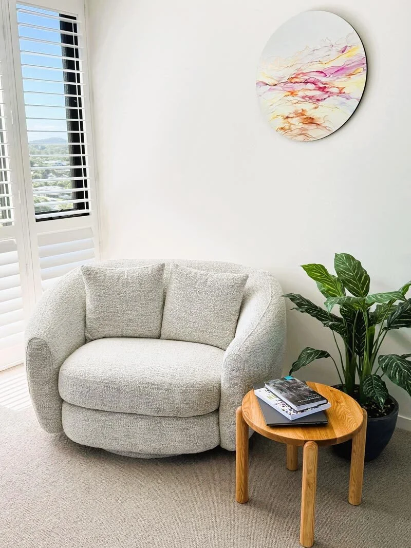 Centred Wellness therapy corner with a light grey upholstered armchair, a small wooden side table with books on it, a large green leafy plant, a circular colourful abstract wall art, and a window with white shutters during daytime.