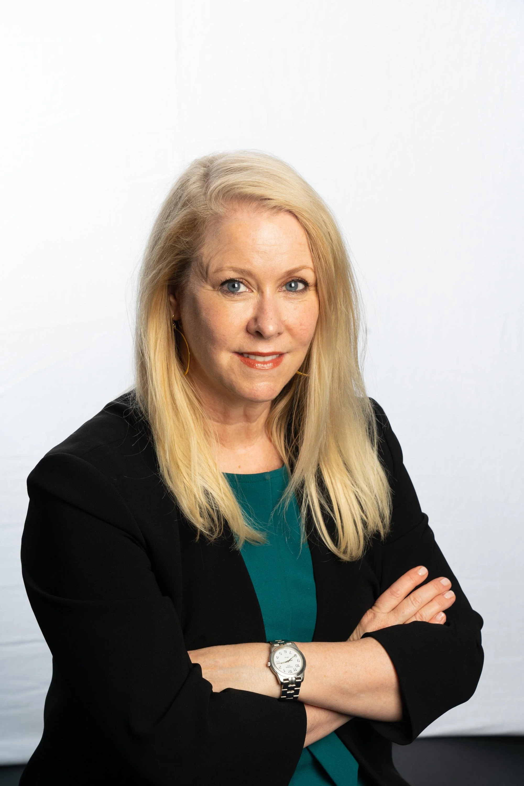 A woman with long blonde hair, wearing a black blazer, teal top, and a watch, standing with crossed arms against a plain white wall.