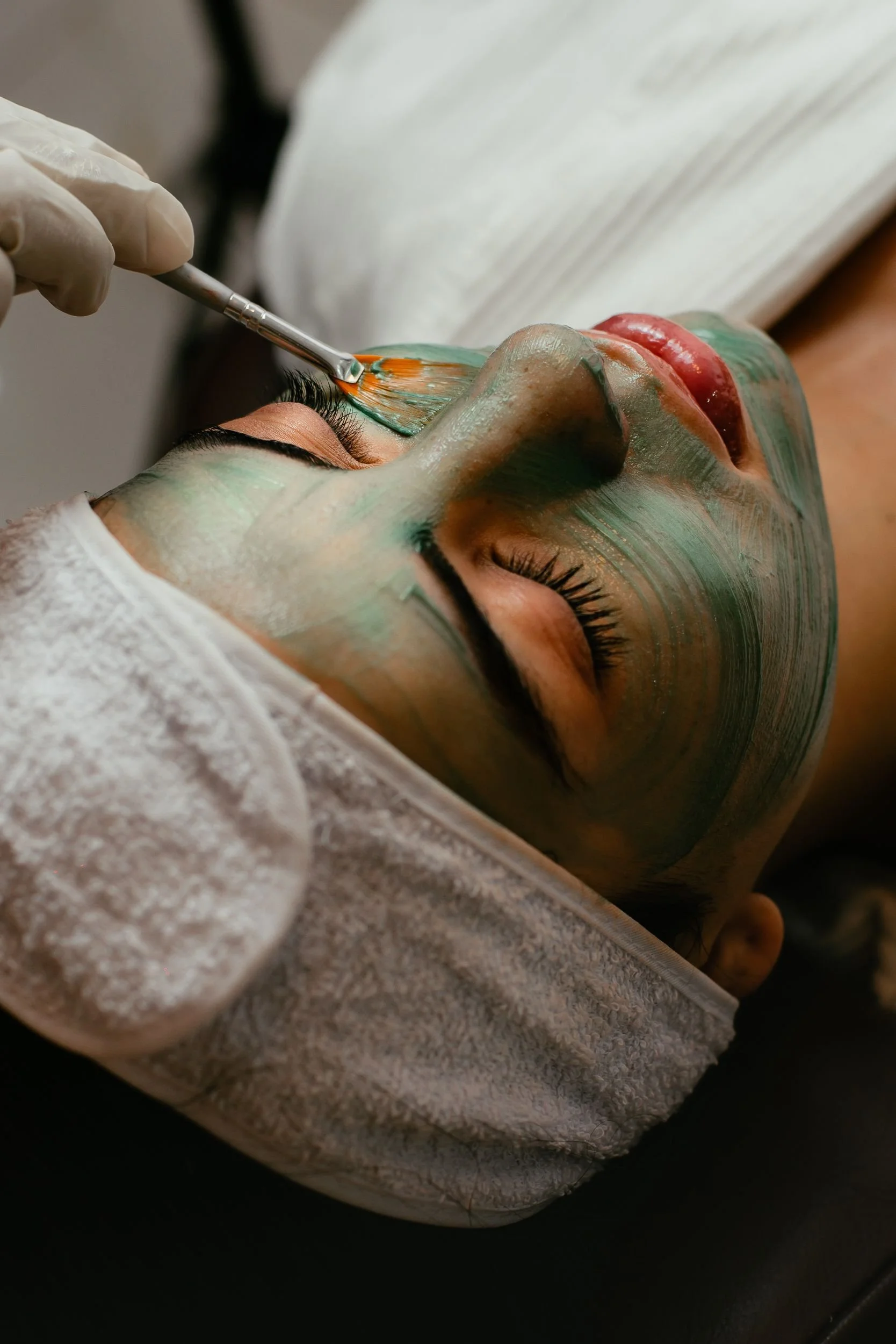A person lying down with their eyes closed, receiving a facial treatment. The person has makeup or face paint on their face, and a professional is applying a facial mask or paint with a brush.