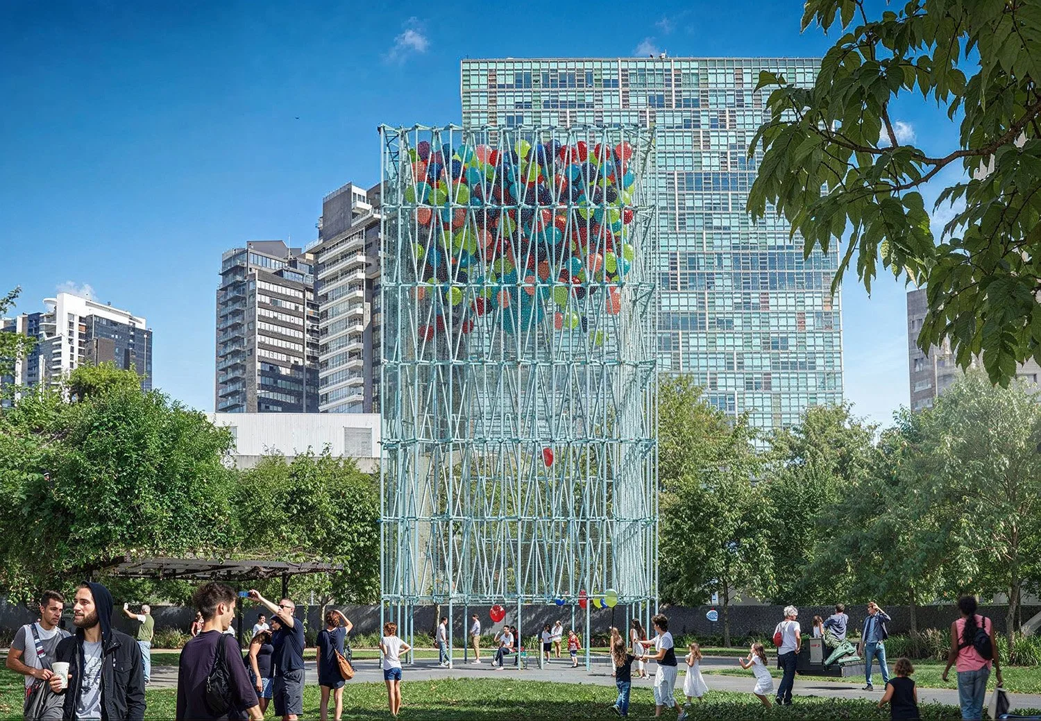 Public park with large climbing structure made of metal pipes and colorful colorful balls at the top. Surrounding trees and city buildings in the background with people walking and children playing.