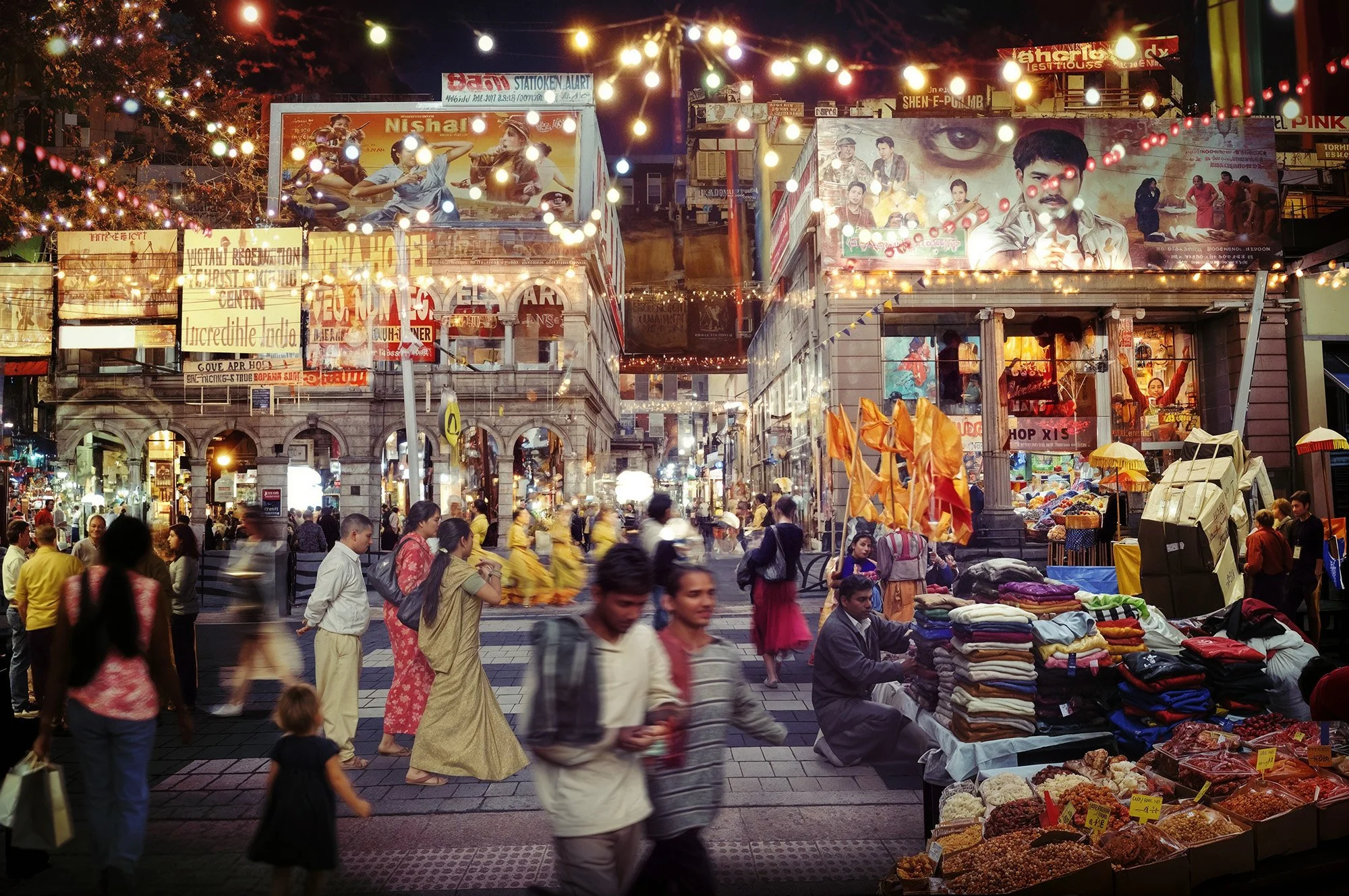 A busy night street market in India with people walking, street vendors selling folded fabrics and dried fruits, illuminated signs, and brightly lit buildings overhead.