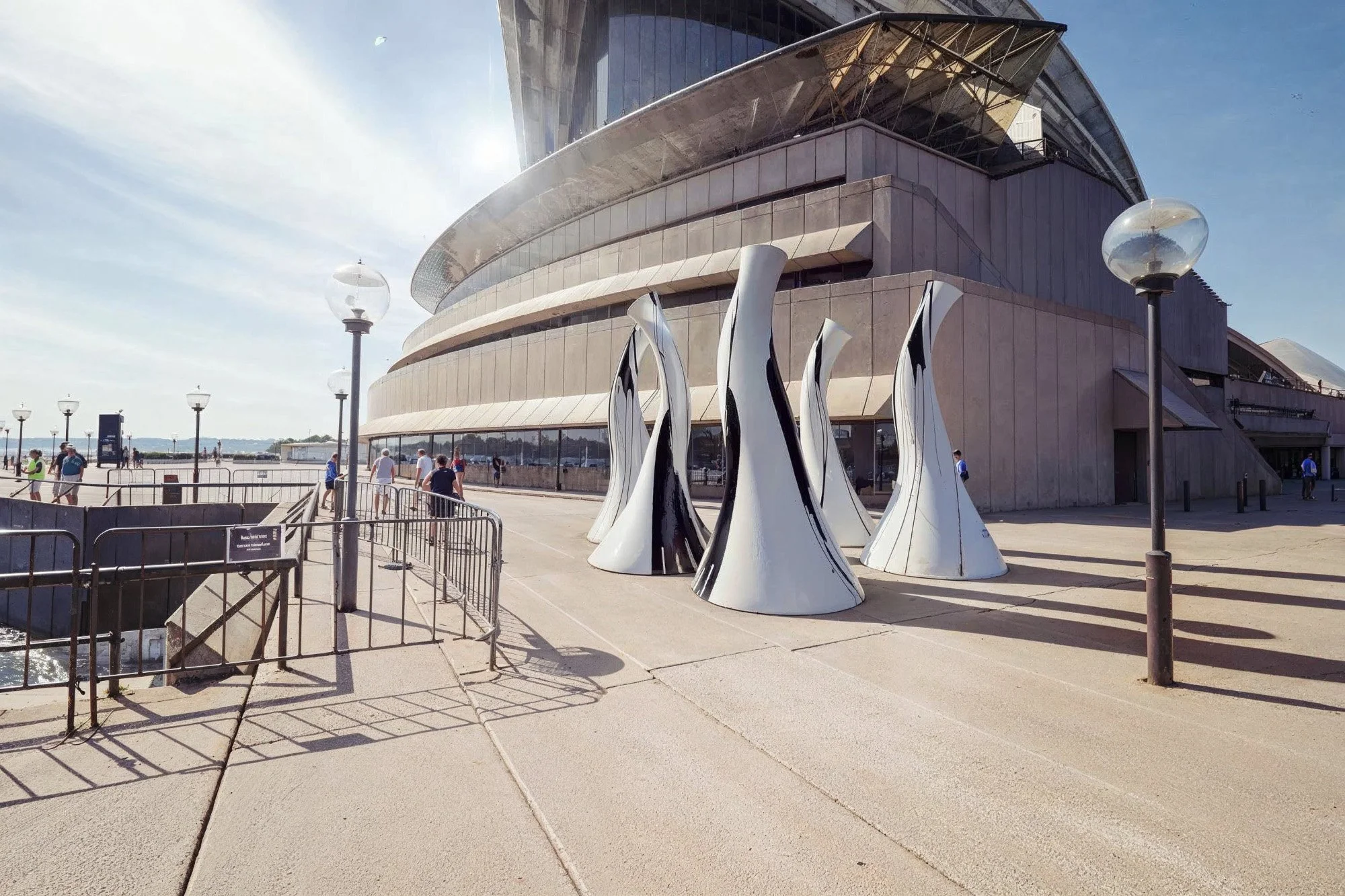 Modern building with curved architecture and glass facade, outdoor sculptures resembling elongated flames or tentacles, lamp posts, and a few people walking outside on a sunny day.