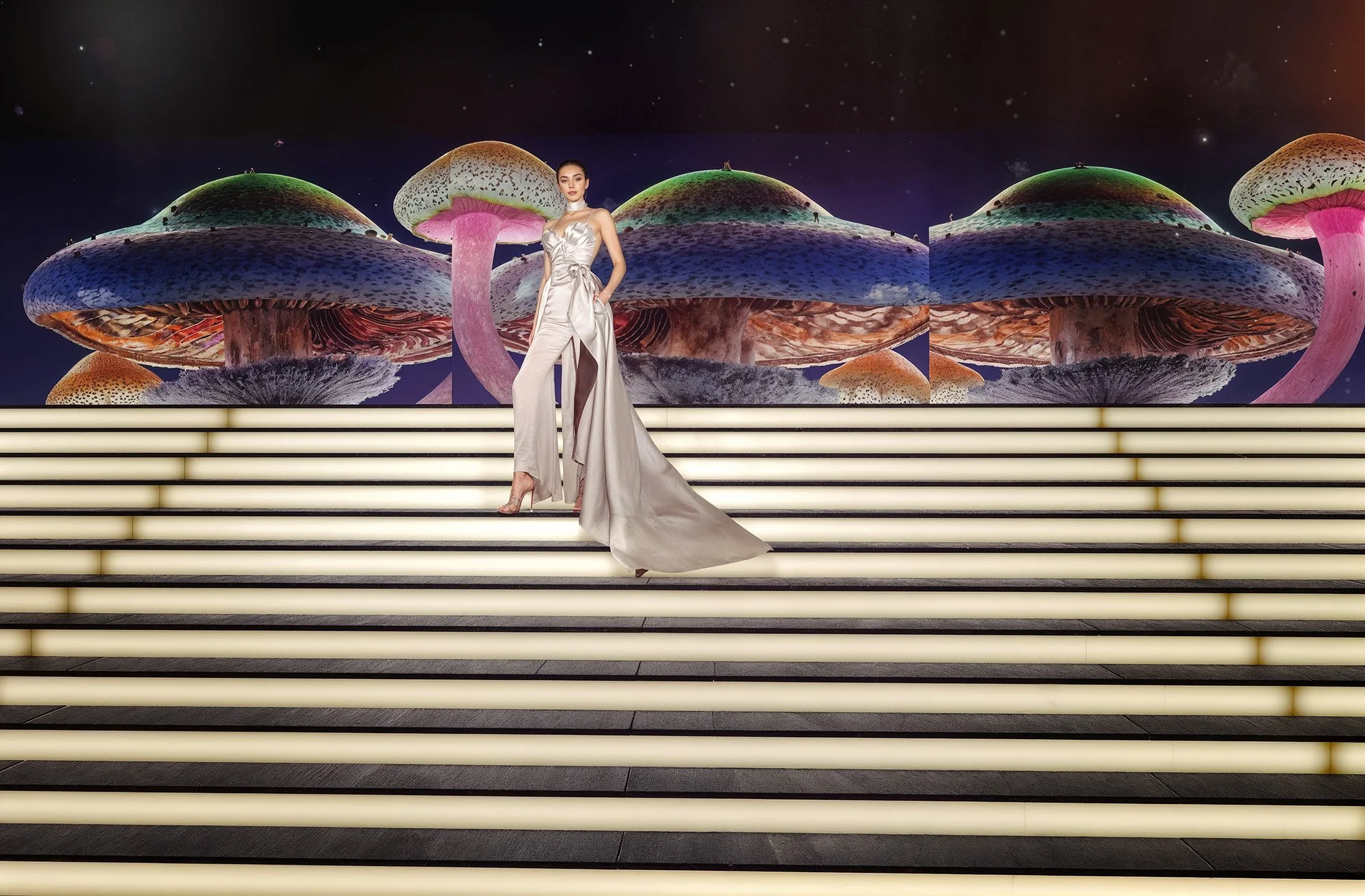 A woman in a white satin gown standing on illuminated steps with giant colorful mushroom sculptures and starry night sky backdrop.