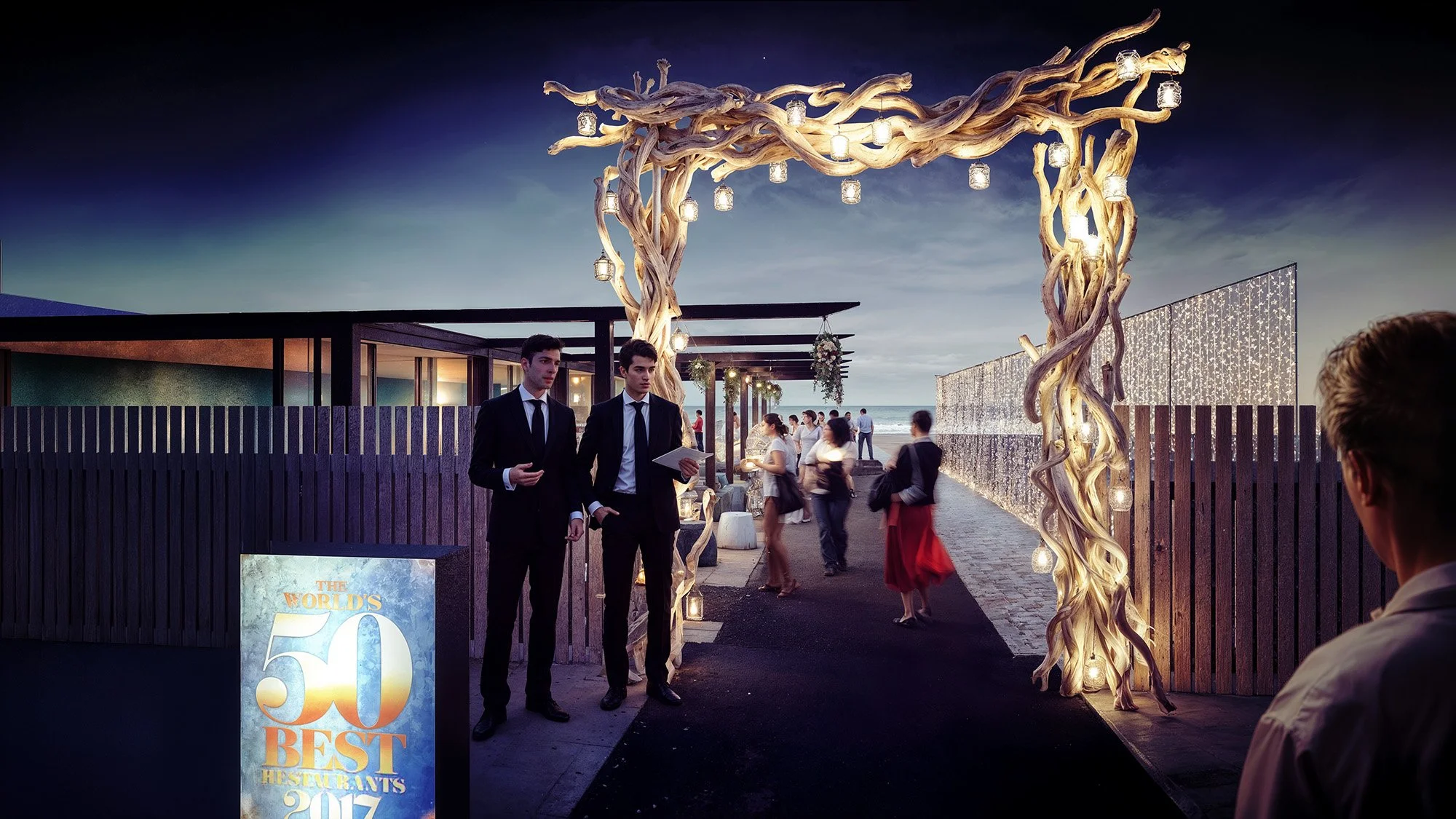 Outdoor evening event with a decorative arch made of twisted tree branches and hanging lights, people mingling in the background, some dressed formally, and a sign indicating 'The World's 50 Best Restaurants 2017' at the entrance.