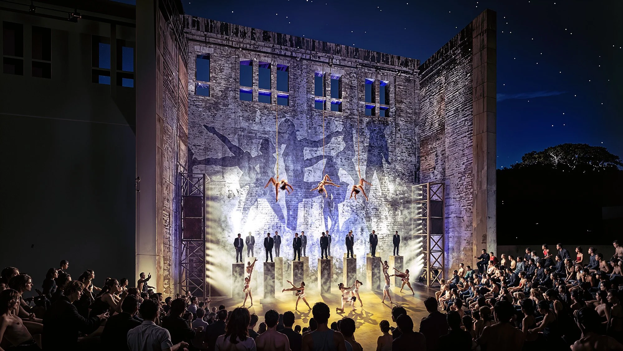 A theater performance with dancers in costumes performing on stage, with a backdrop depicting ballet dancers. The stage is lit with bright lights, and an audience watches outdoors at night under a starry sky.