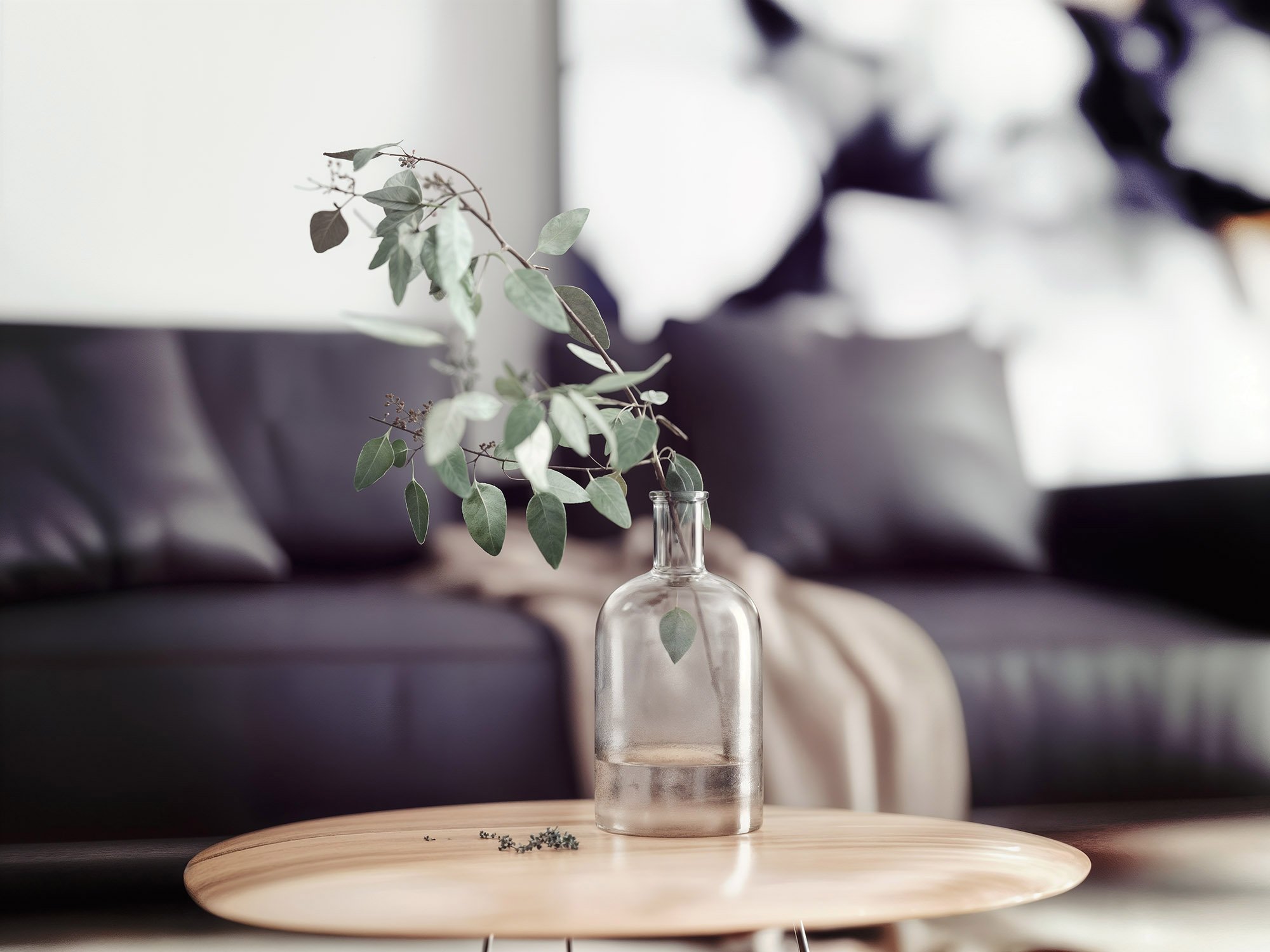 A clear glass bottle with a sprig of greenery inside, placed on a round wooden table in a living room with black sofa and pillows.