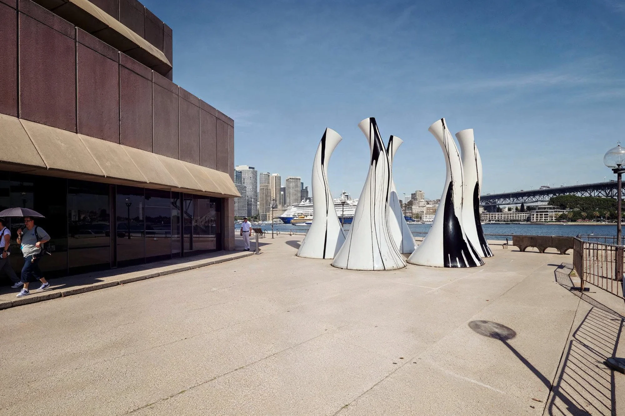 Sculpture installation of white and black cone-shaped structures near a waterfront, with a city skyline and sailboats in the background.