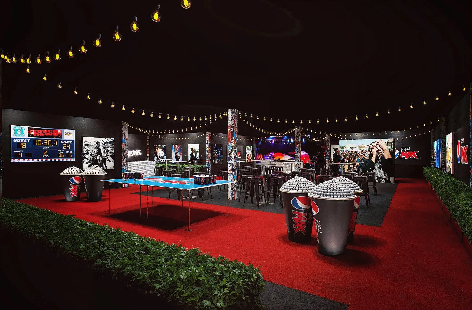 An indoor event space designed like a sports bar with a video scoreboard, ping pong table, large bowls of golf balls, and multiple TV screens showing sports and Pepsi Max branding. The ceiling is decorated with string lights, and there are tables and