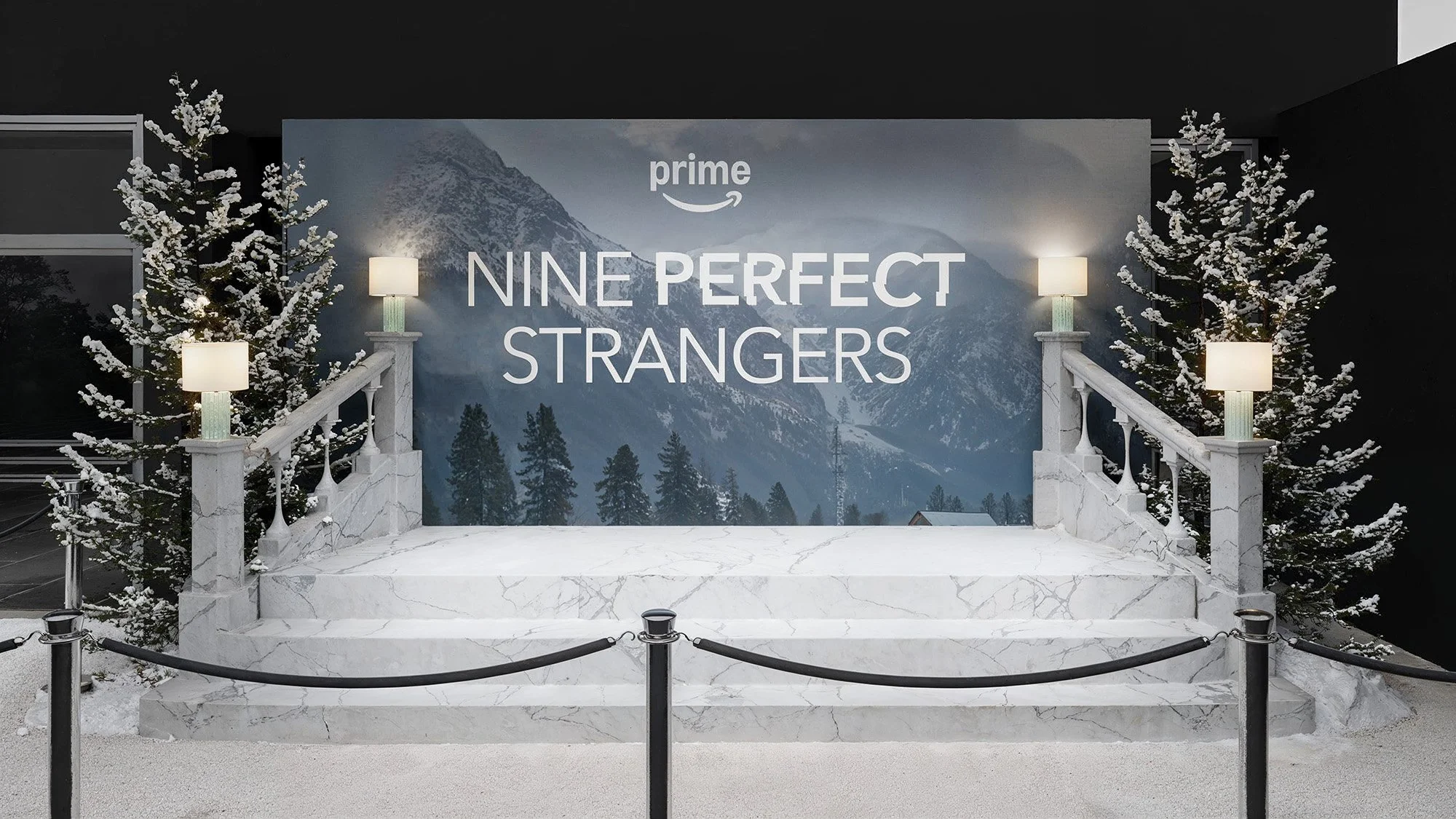Stage with a marble platform, white street lamps, snow-covered trees, and a mountain backdrop, advertising the TV show 'Nine Perfect Strangers' on Amazon Prime.
