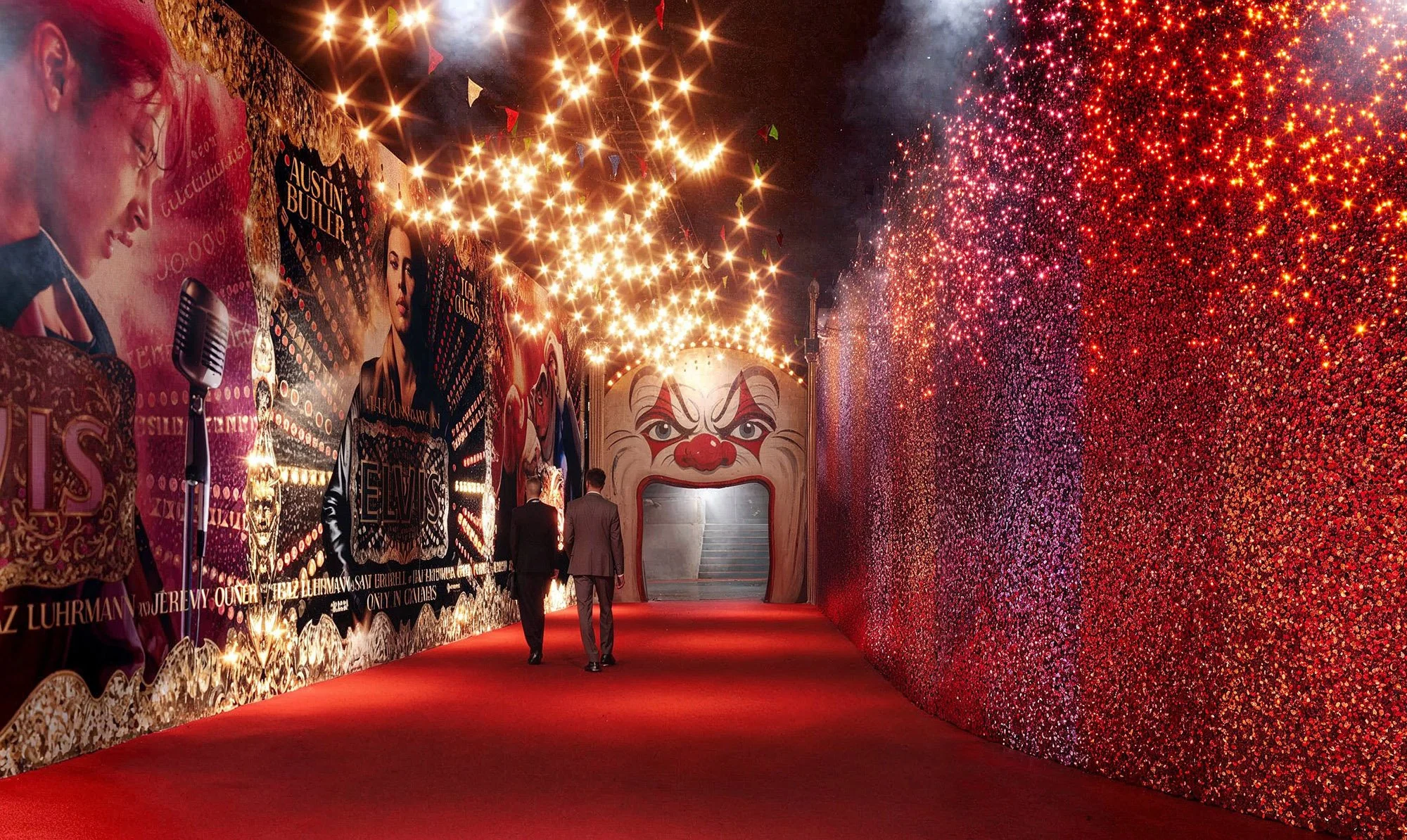 Two men in suits walking down a red carpet tunnel decorated with singer posters on one side and a large clown face with an open mouth at the end. The ceiling has string lights and fireworks in the background.