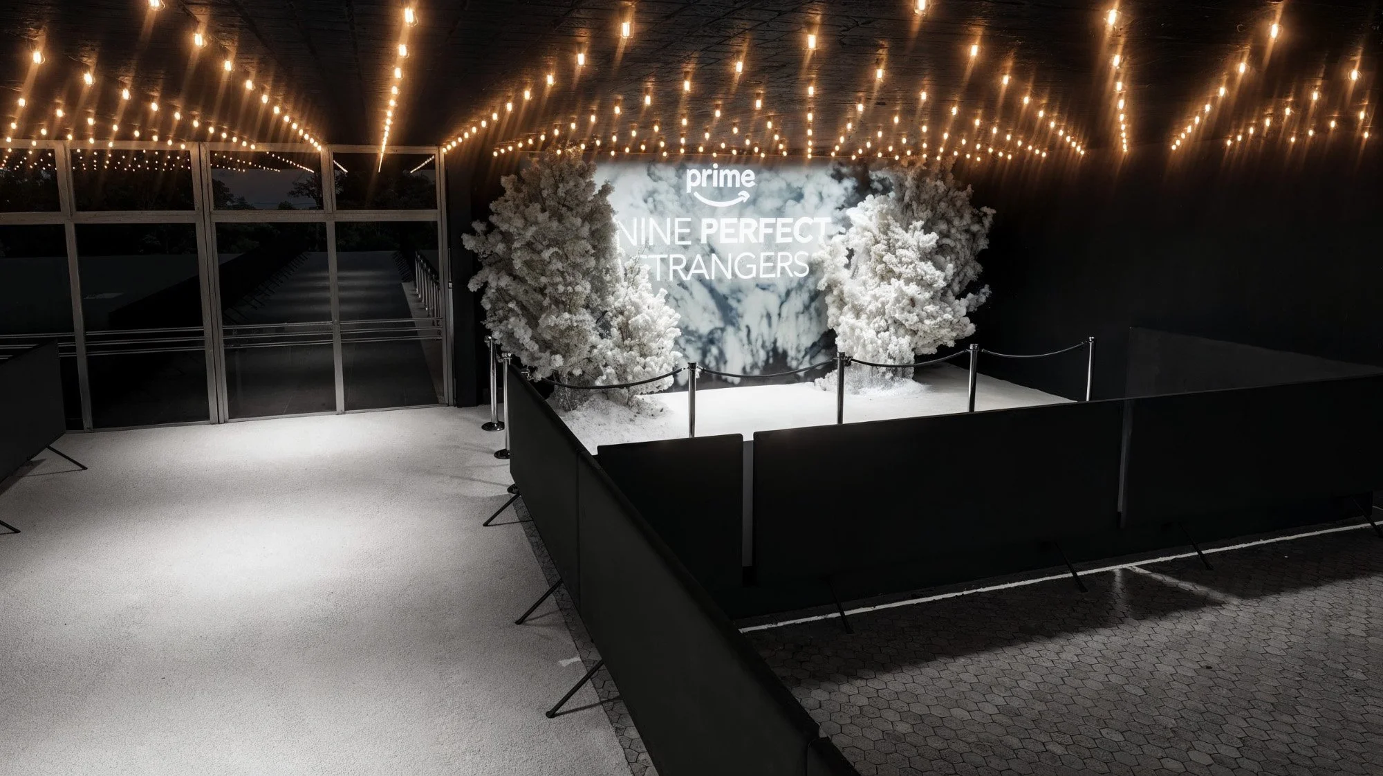 Indoor event space with string lights on ceiling, decorated with white snowy trees in front of a large screen displaying 'prime NINE PERFECT STRANGERS,' surrounded by black barriers.