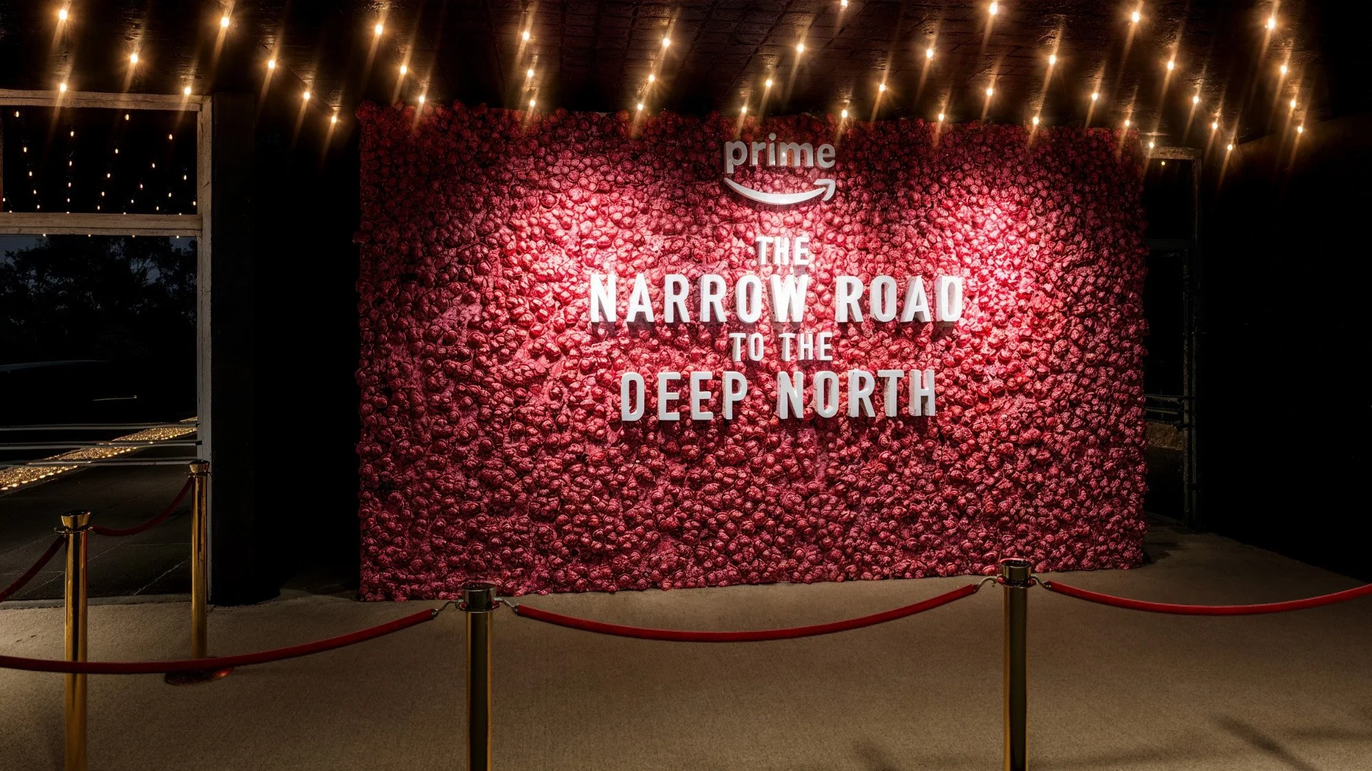 A wall covered with pink roses and white letters spelling out "The Narrow Road to the Deep North" with the Amazon Prime logo above, inside a decorated room with string lights and a red velvet rope barrier.