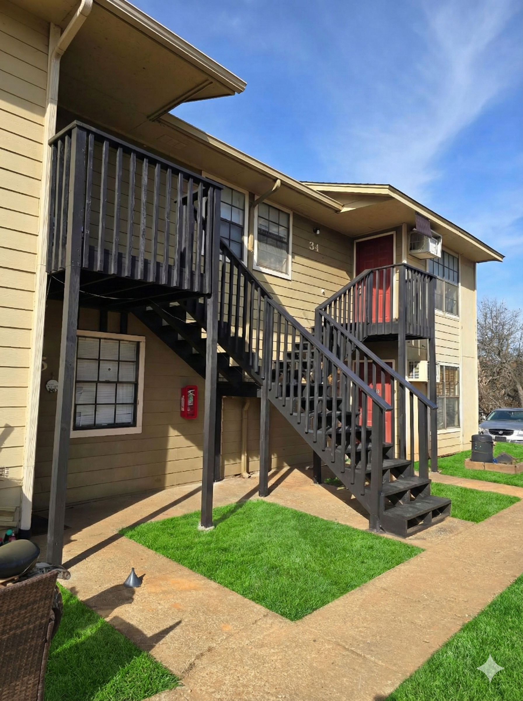 Exterior view of an apartment building with a staircase leading to the second floor, beige siding, red doors, and a small patch of green grass on a sunny day.
