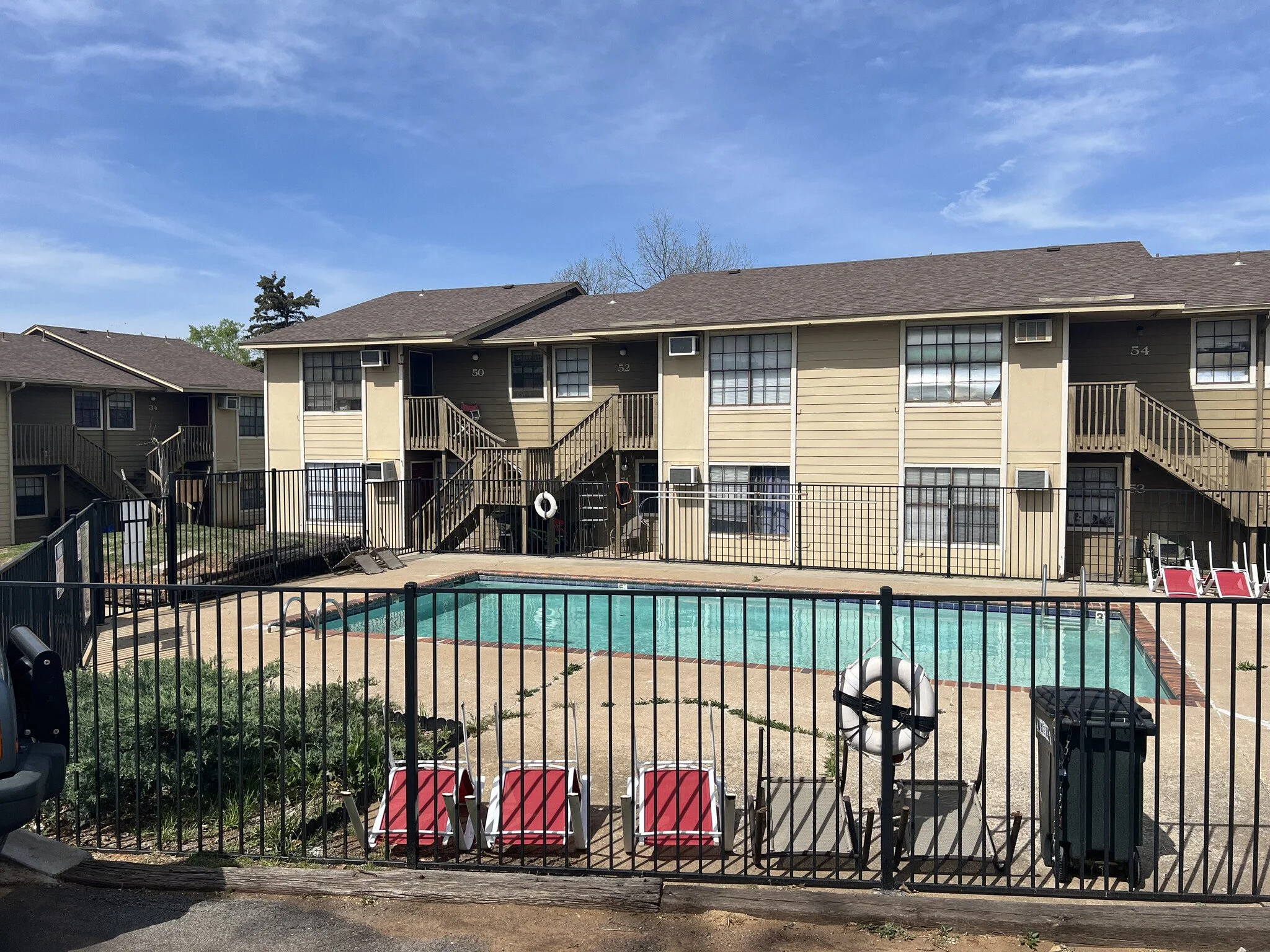 Apartment complex with swimming pool, pool chairs, and a fence around the pool area, with two-story buildings in the background.