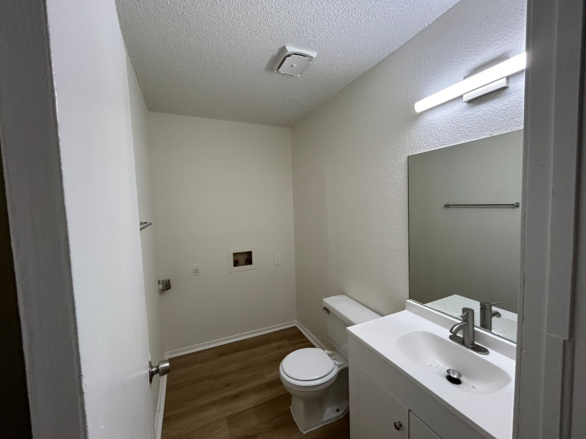 A small, empty bathroom with white walls, a toilet, a white sink with a mirror above, a light fixture, and a wood-look floor.