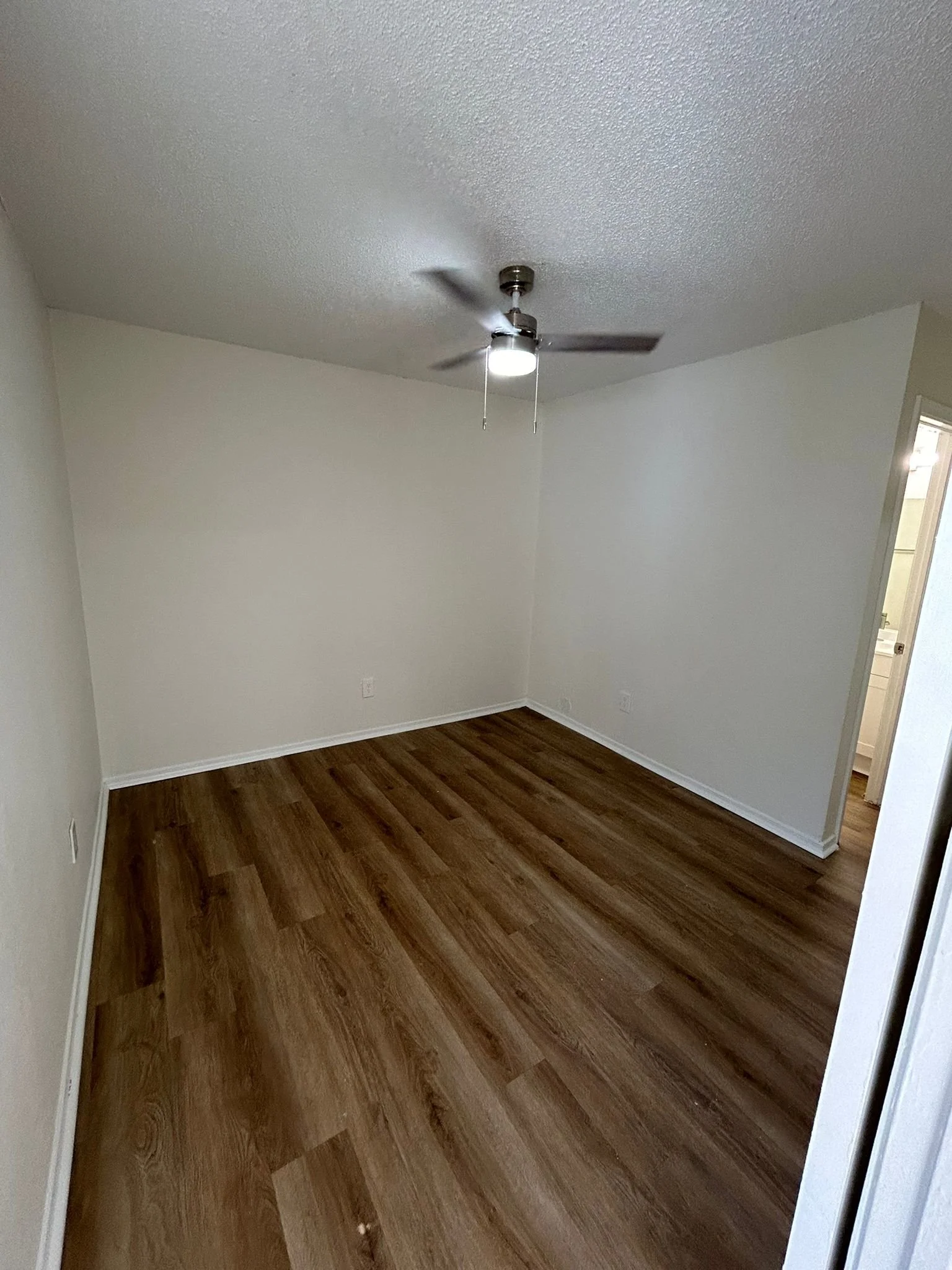 Empty room with white walls, wood laminate flooring, and a ceiling fan with lights turned on.