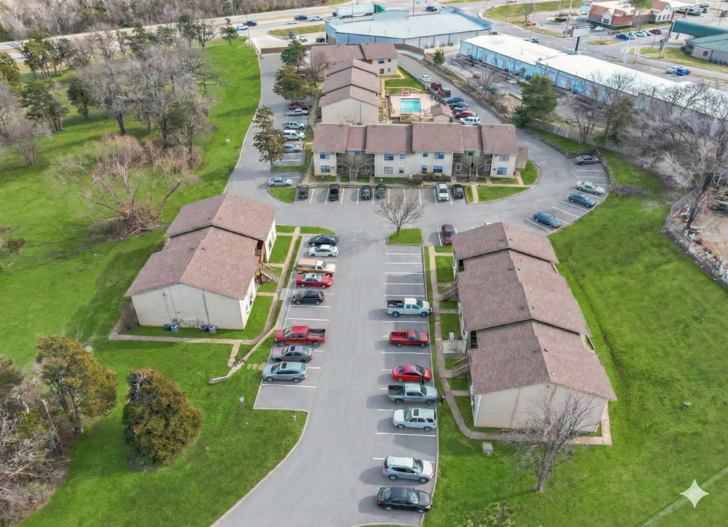 Aerial view of a residential apartment complex with multiple buildings, parking lots with cars, surrounding green lawns, and trees. There is a swimming pool visible in the central area.