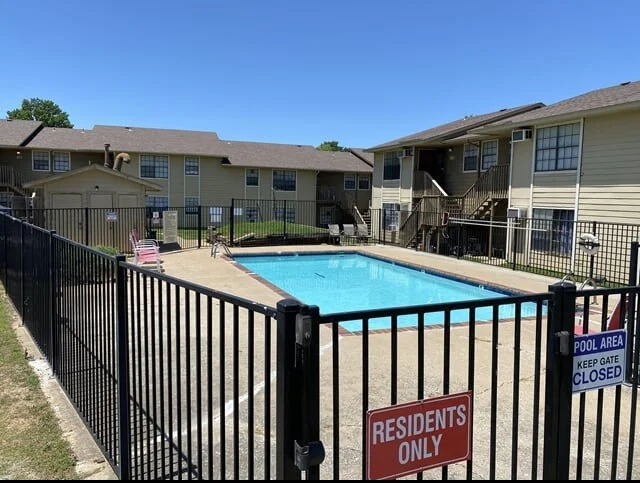 Residential apartment complex with a fenced swimming pool area, lounge chairs, and a clear blue sky.