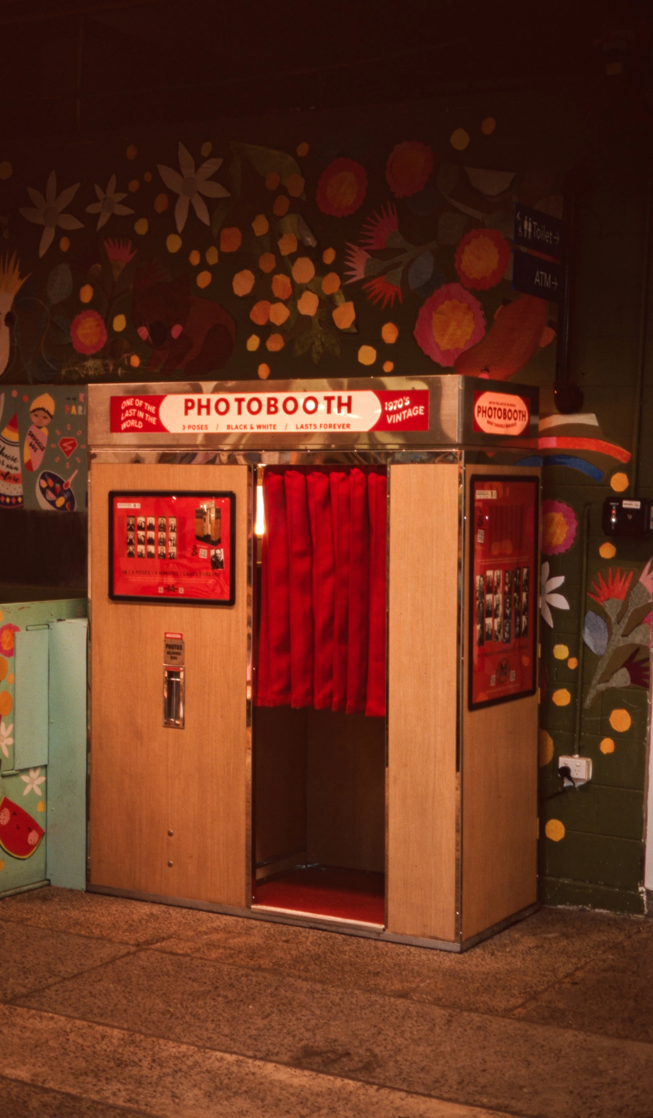 film 35mm colour photo of a wood grain 1970's vintage analogue photobooth with red curtin and painted floral wall behind in warm tones