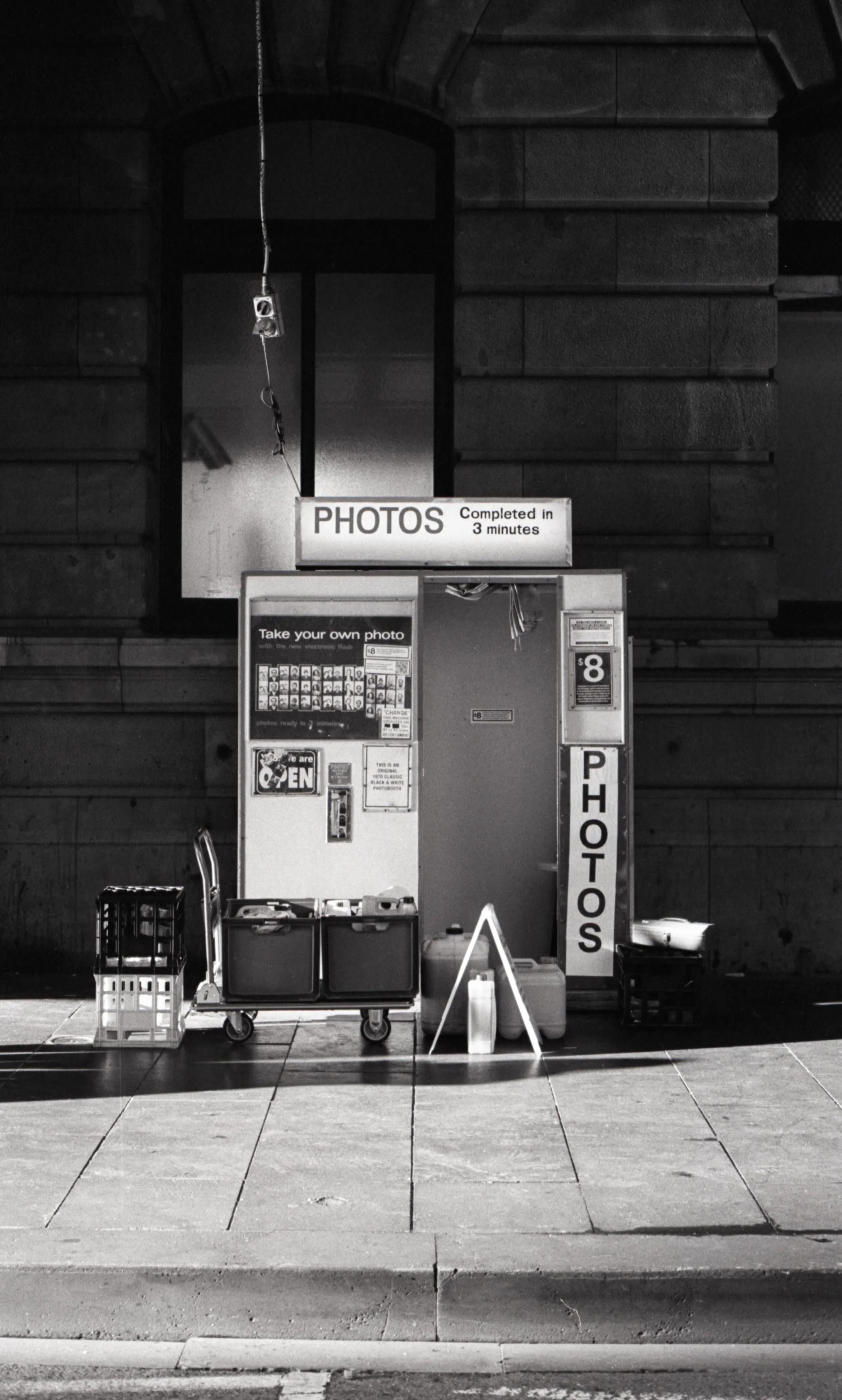 A black and white photo of a self-service photo booth on a city sidewalk, with a sign indicating photos are completed in three minutes and a menu for taking your own photo.