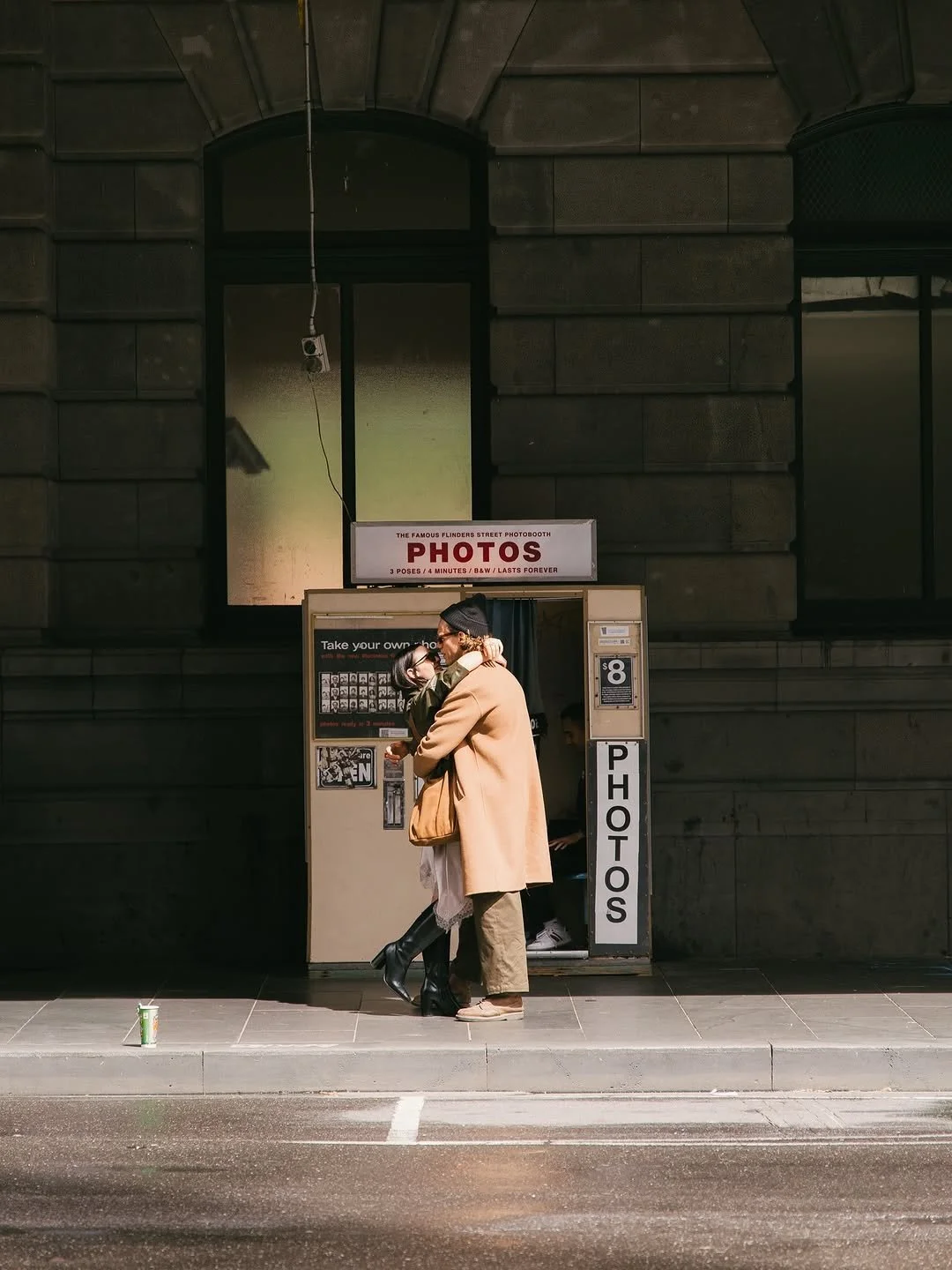 A couple embraces in front of a street photo booth, with a sign that says 'PHOTOS'. The photo booth is on a city sidewalk, with a beverage cup on the ground nearby. The pair is illuminated by sunlight against a dark building facade.