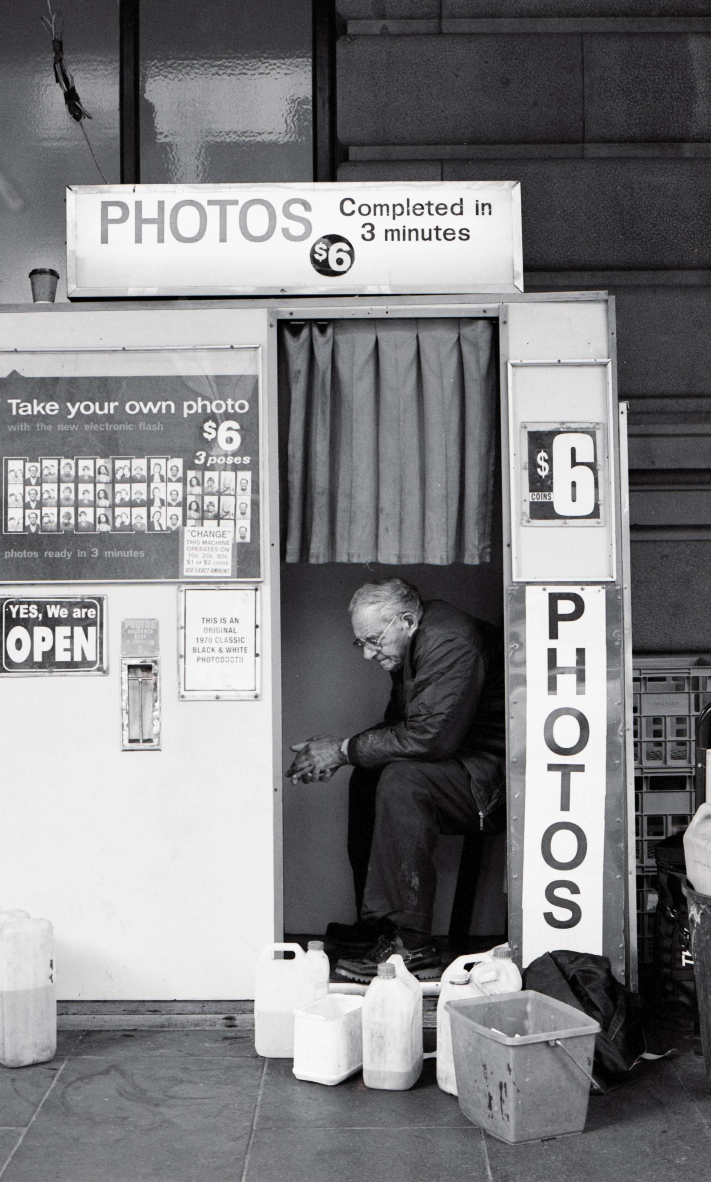 A man sitting in a photo booth on a city sidewalk, surrounded by gallon jugs and a plastic container, with signs indicating photo prices and service details.