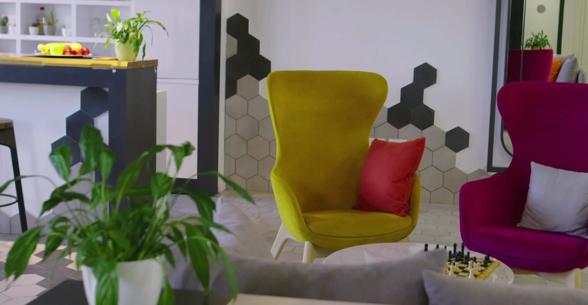 Colorful living room with two high-back chairs, one yellow and one magenta, each with a throw pillow; a small table with a chessboard, a potted plant in the foreground, and honeycomb-shaped wall art.