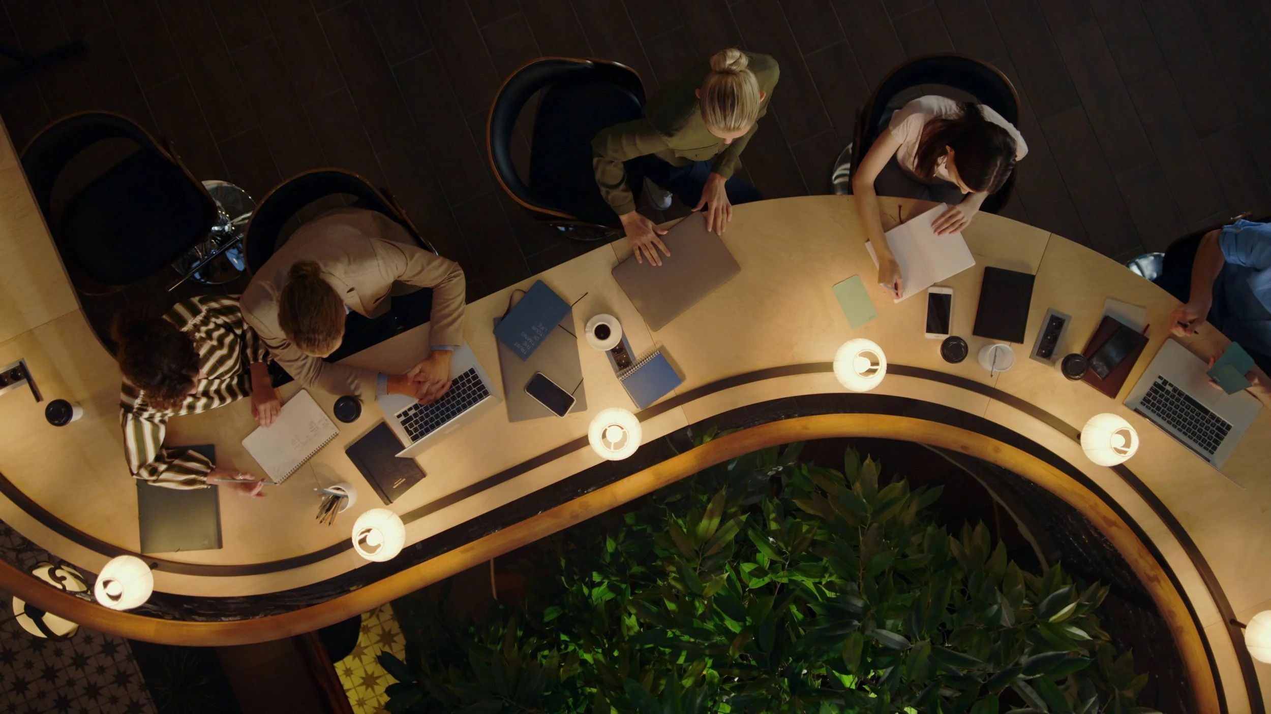 Top-down view of five people working at a curved wooden conference table with laptops, notebooks, phones, and coffee cups, surrounded by ceiling lights and a green plant.