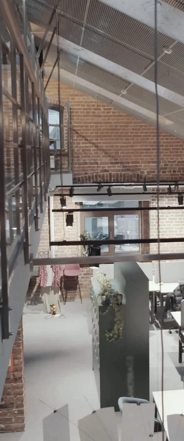 Interior view of a modern industrial-style office space with exposed brick walls, metal railings, a lofted ceiling, and large windows.