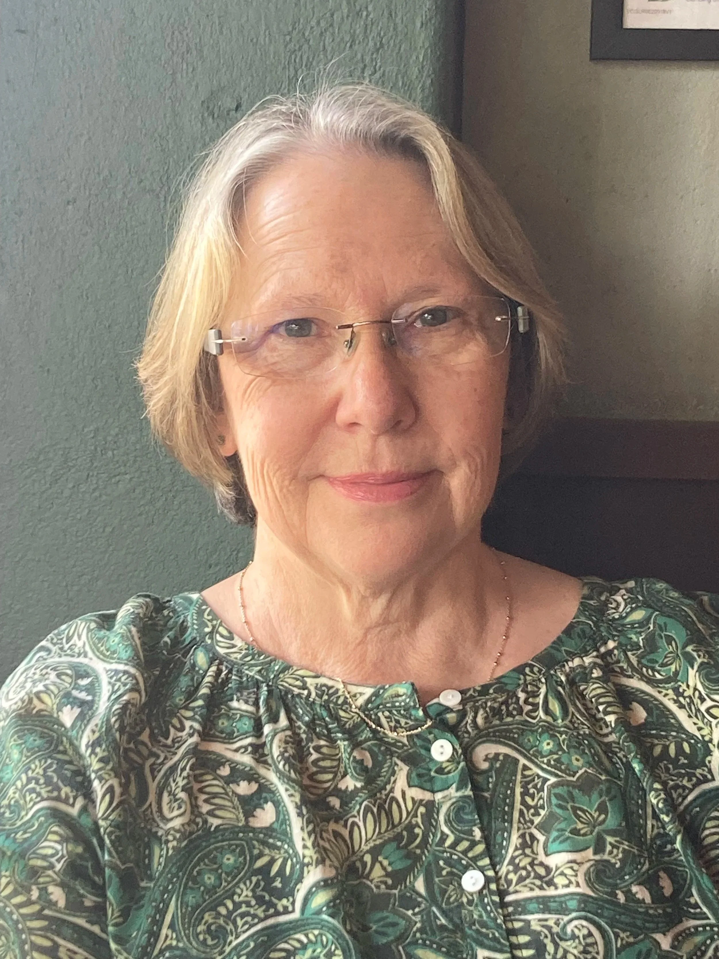 A middle-aged woman with short, light-colored hair wearing glasses and a paisley-patterned blouse, sitting indoors in front of a green wall.