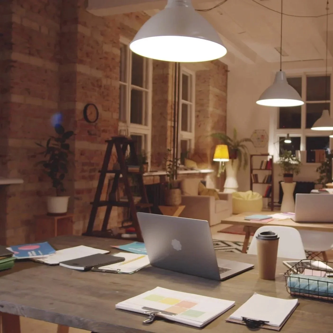 An illuminated office or workspace with a large wooden table, laptops, notebooks, a coffee cup, and stationery. The background shows brick walls, large windows, potted plants, and modern hanging lights.