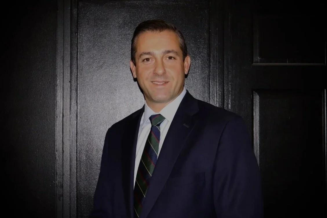 A man in a dark suit with a striped tie, standing against a black wood-paneled background, smiling at the camera.