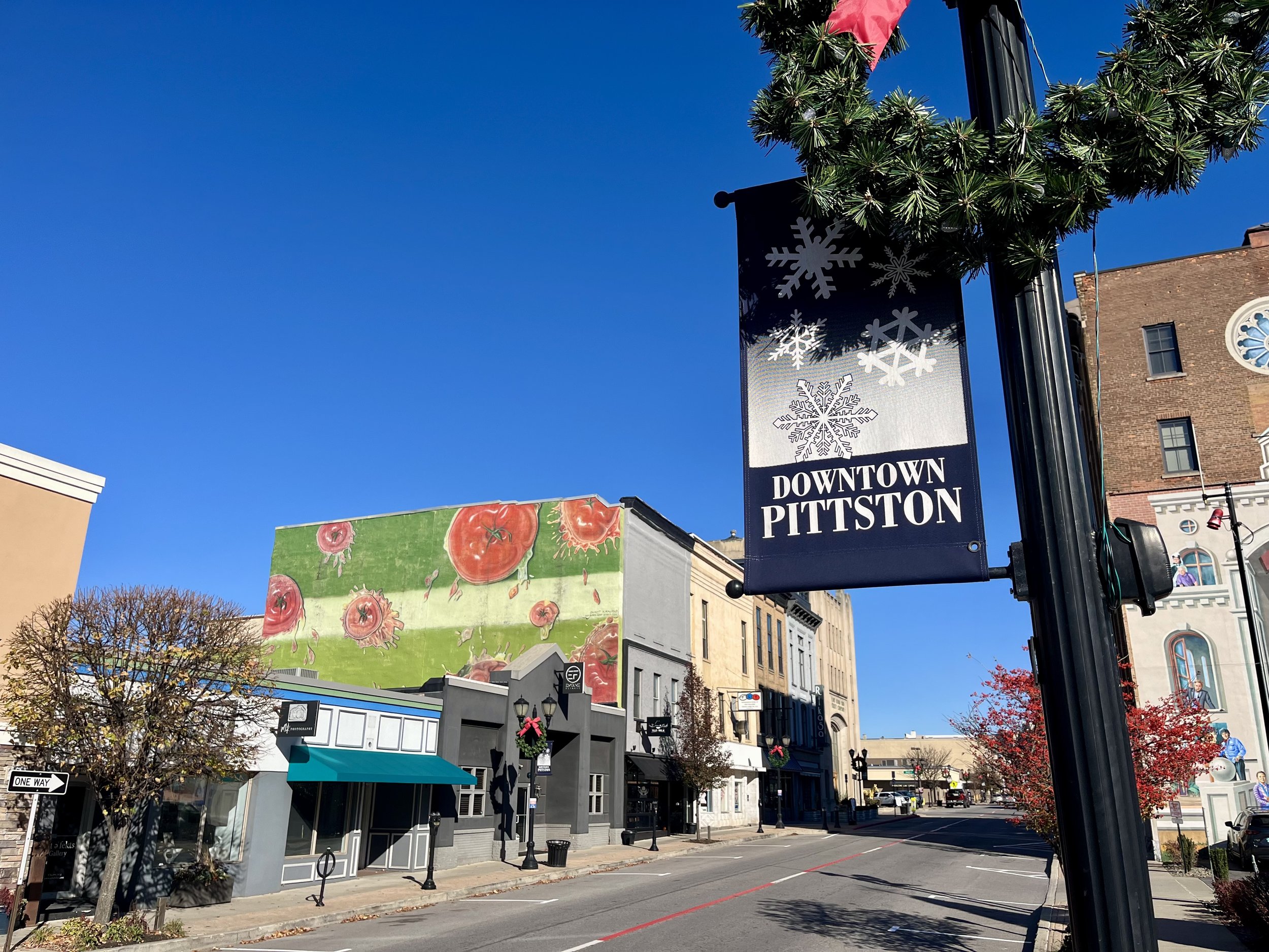Downtown Pittston decorated for the holidays, featuring signs with snowflakes, a large tomato mural on a building, and buildings with festive decorations on the street.