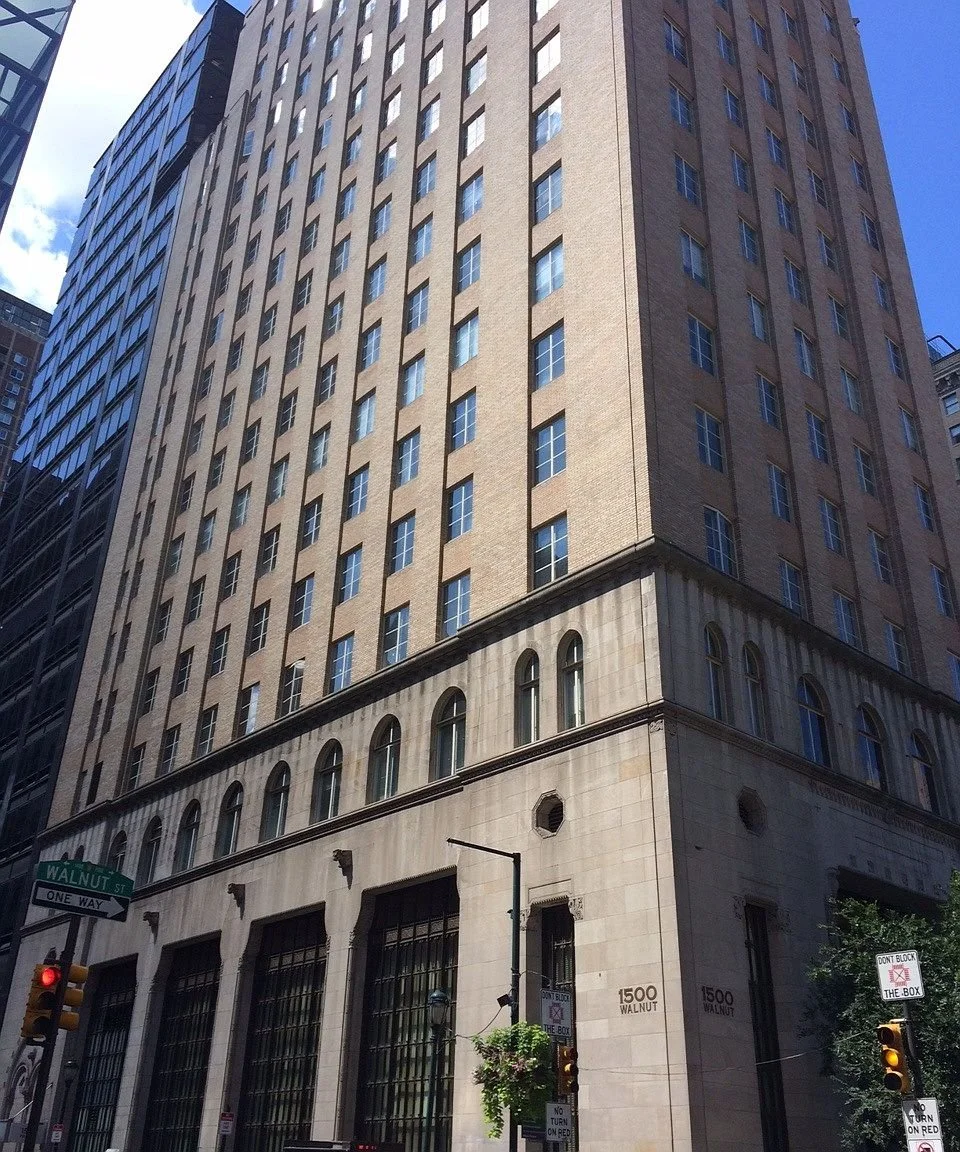 A tall, multi-story building with brick and stone facades, large windows, and street signs at an urban intersection.