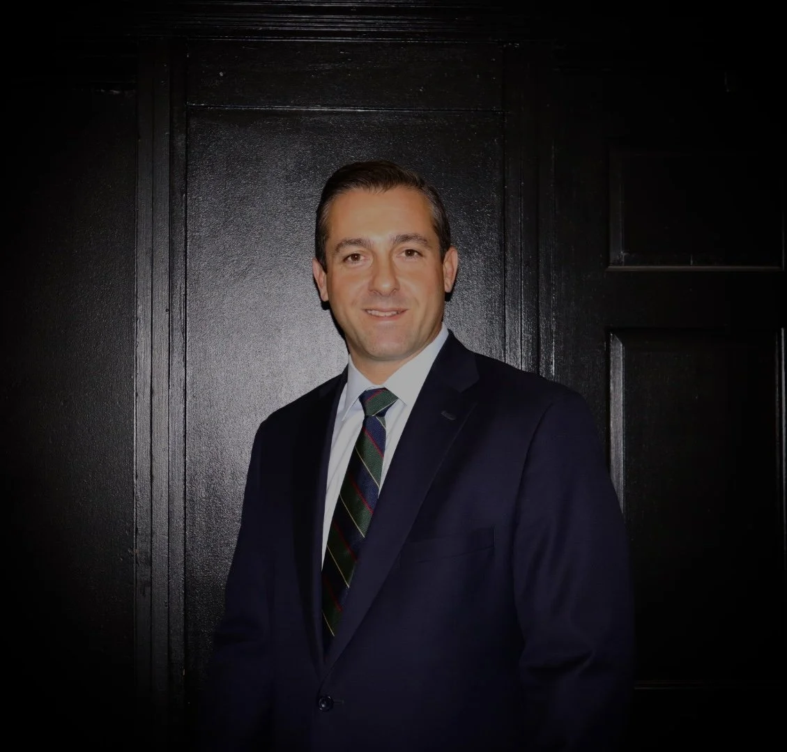 A man wearing a dark suit, white shirt, and striped tie, standing in front of a dark background, smiling at the camera.